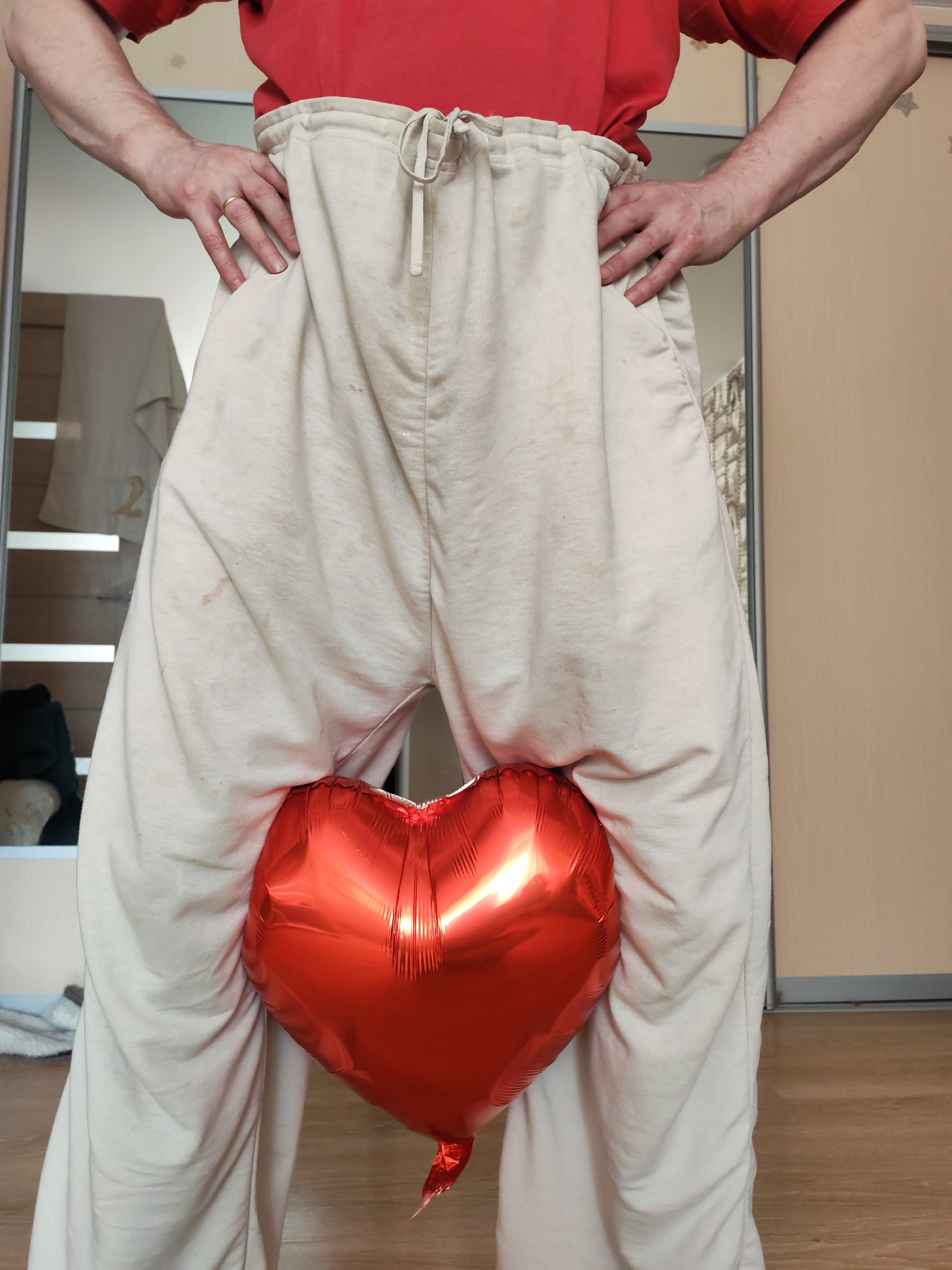An individual is depicted indoors, likely in the city of Chaikovsky, Russia, standing with hands placed on their hips. The person is wearing a red short-sleeved t-shirt and light beige drawstring sweatpants, which display several faint light brown stains. A plain gold band is visible on the ring finger of the left hand. Positioned between the person's legs, at the crotch level, is a reflective, inflated red heart-shaped foil balloon. The background consists of a large mirror reflecting a light-colored garment and an interior space, alongside a light brown wall and a light wood-patterned floor.
