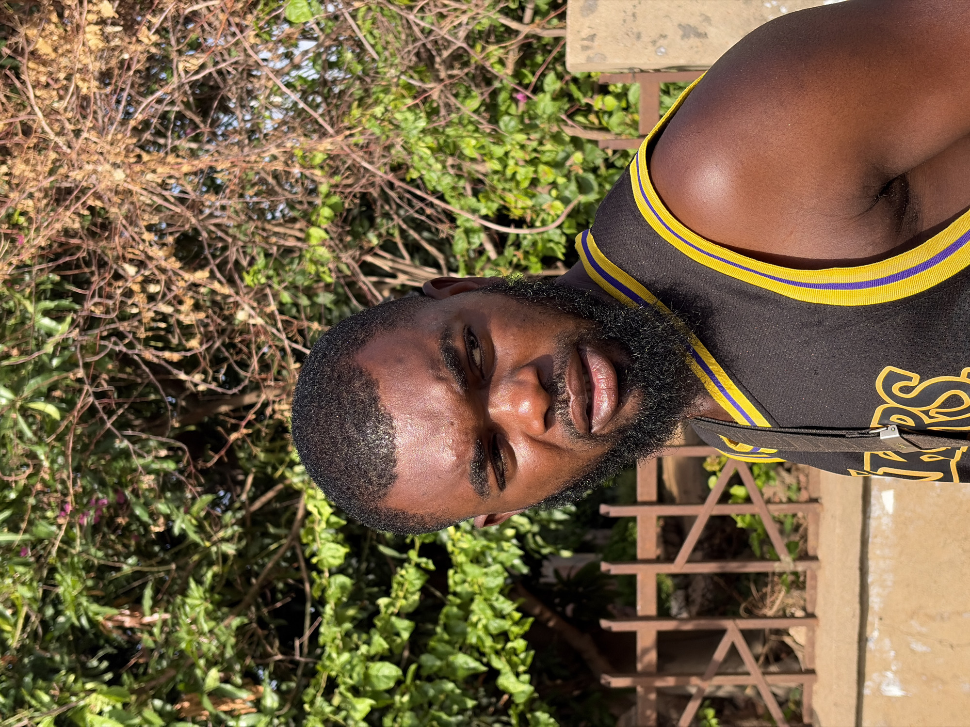 A medium outdoor shot shows a Black male wearing a black and gold basketball jersey. He is centered and faces the camera directly. The man has short hair, a trimmed beard, and is positioned outdoors. Behind him, there is greenery, including a leafy bush and some bare branches. A metal railing is also visible in the background, along with a concrete wall. The location is possibly near Jos, Nigeria, given the vegetation and light conditions. The image is taken during the daytime in natural sunlight.