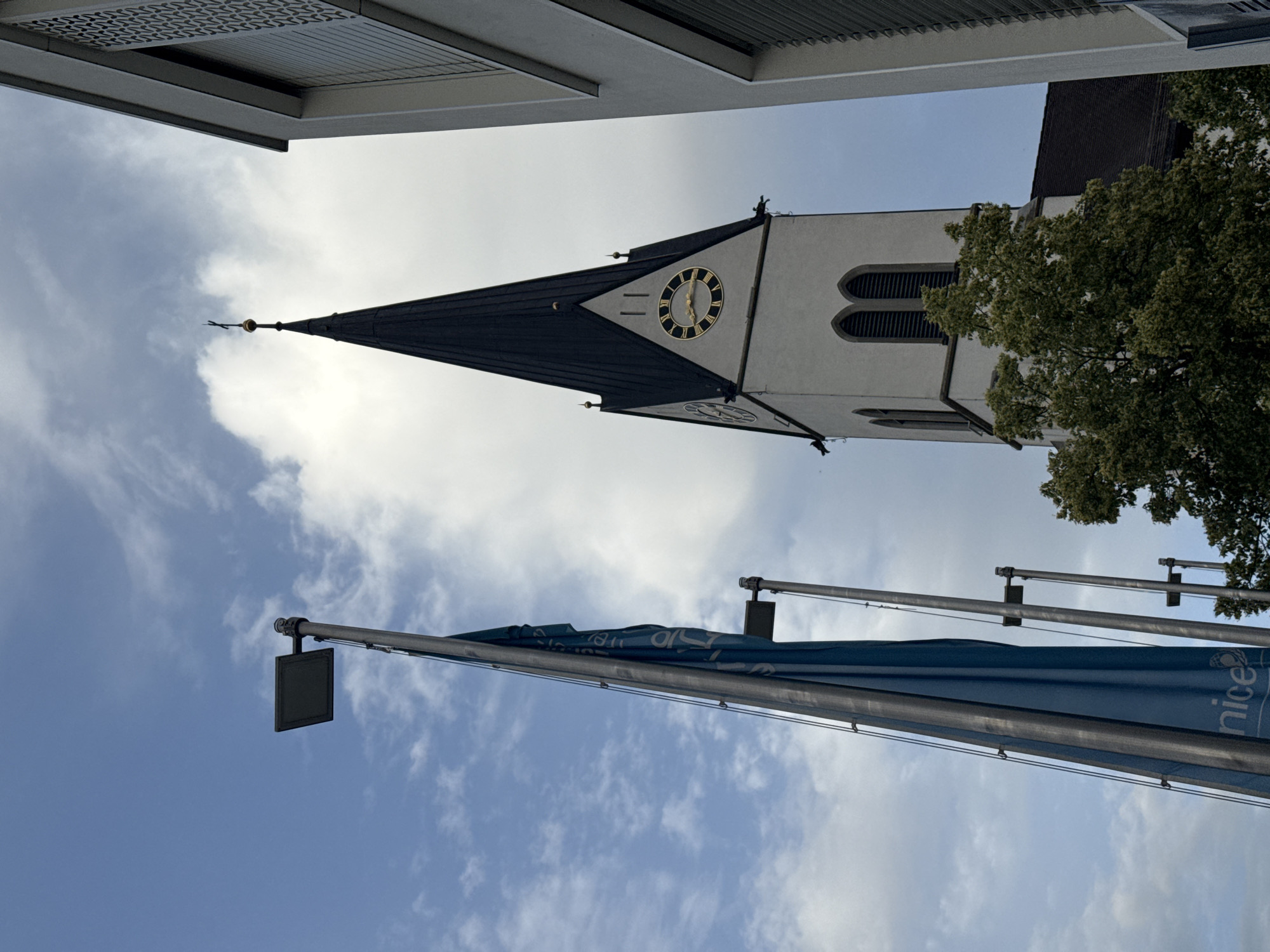 The photograph captures an upward view of a church tower in Nendeln, Liechtenstein. The tower, primarily white, features a dark, pointed spire topped with a cross. A prominent clock face with Roman numerals is visible on the tower. To the left of the tower, a tall flagpole with a large, blue flag is present. The flag appears to have white lettering and a logo, potentially associated with UNICEF. A modern building with horizontal window blinds is situated to the right of the church tower. The sky is partly cloudy with patches of blue visible. Green trees are partially obscuring the lower section of the church.