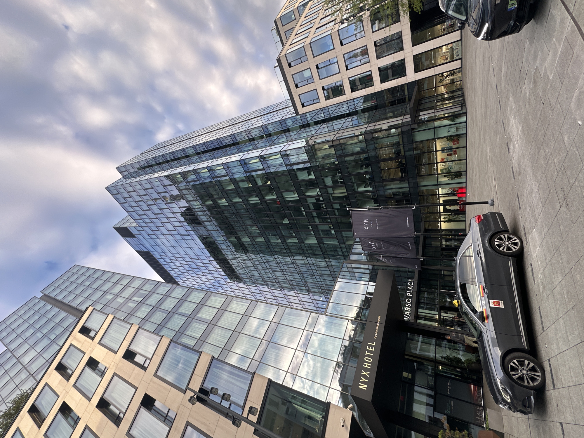 The image shows the facade of the NYX Hotel by Leonardo Hotels, located in Warsaw Place, Warsaw, Poland. A dark grey sedan, identified as a taxi by markings on its door and a yellow light on the roof, is parked in front of the hotel entrance. Two large black banners with the NYX Hotel logo are displayed on flagpoles. The hotel is situated within a complex of modern glass and concrete buildings, with one prominent skyscraper reflecting the cloudy sky. The foreground is a paved plaza with light grey square tiles. The sky is overcast with dramatic clouds.