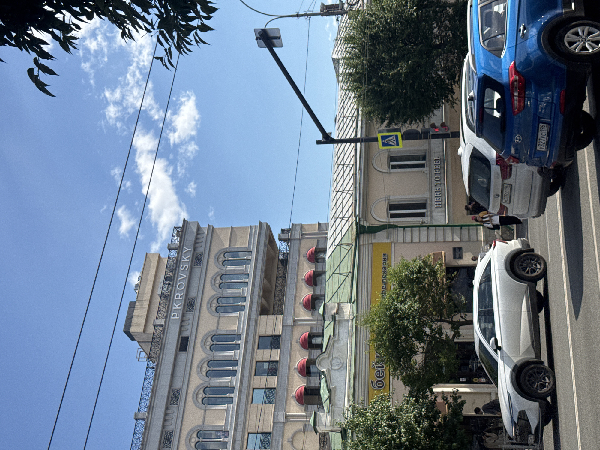 This is a downward-facing, street-level shot taken in Yekaterinburg, Russia. The sky is bright blue with scattered white clouds, suggesting fair weather. The time of day appears to be daytime, likely late morning or early afternoon due to the sunlight casting shadows.
The scene features several cars parked or driving on a paved street. A prominent blue SUV is on the right, followed by a white SUV. A white sedan is also visible, with its rear end facing the viewer. There are a few people visible, some near the cars and others walking on the sidewalk.
The background is dominated by buildings. On the left, a tall, ornate building with "PKROVSKY" written vertically on its facade is visible. This building has arched windows and decorative elements. To the right of this is a lighter-colored building with a green awning and a yellow sign that appears to have Russian text on it, including "пекарня" which means "bakery." Another building behind it has the text "HERE TO FEEL" written horizontally. Street signs are also present, including a pedestrian crossing sign. Overhead wires crisscross the scene, and a tree with green foliage is visible on the right side, partially obscuring the buildings.