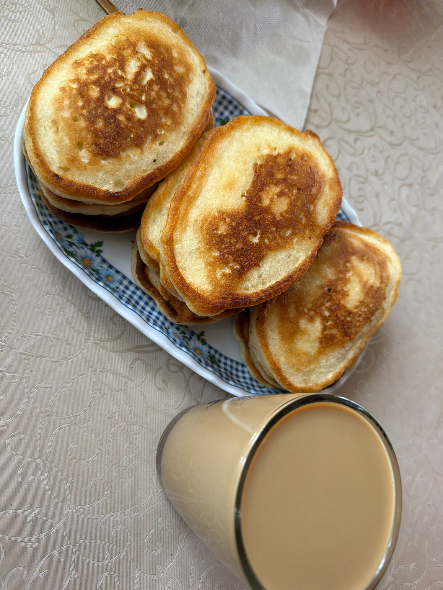 A stack of golden-brown pancakes sits on a white oval plate with a blue and white checkered rim decorated with small blue flowers. To the right of the plate is a tall glass filled with a light brown beverage, likely coffee or tea with milk. The plate and glass rest on a light beige tablecloth with a subtle embossed pattern of swirling vines. The image is taken from a top-down perspective. The location in Teterevo, Russia, is not discernible from the visual information provided. There are no people or actions depicted in the scene.