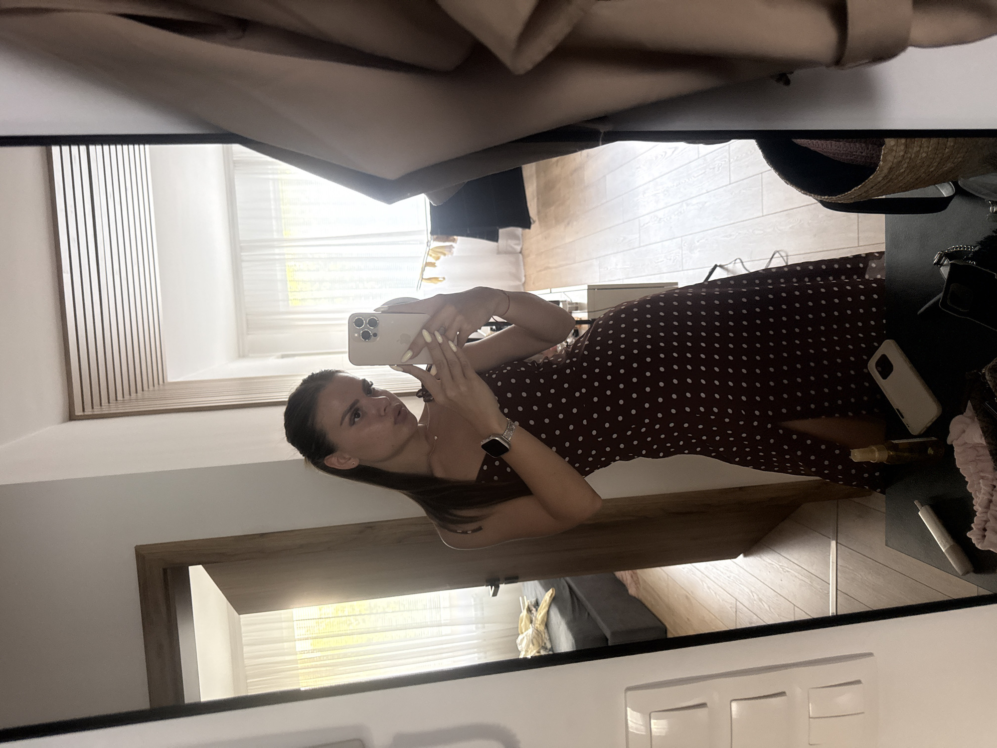 In a well-lit room, a young woman stands before a full-length mirror, taking a selfie with her smartphone. She is wearing a dark, polka-dotted dress with a thigh-high slit. Her hair is dark and pulled back, and she has on a watch. The room appears to be a bedroom or dressing area, with a bed visible in the background through an open doorway. The floor is hardwood, and there are sheer curtains on a window. On a table in the foreground, several objects are present, including a phone, a small bottle, and a woven basket. A neutral-colored coat hangs on the right side of the frame. The scene suggests a personal, indoor setting in Plovdiv, Bulgaria.