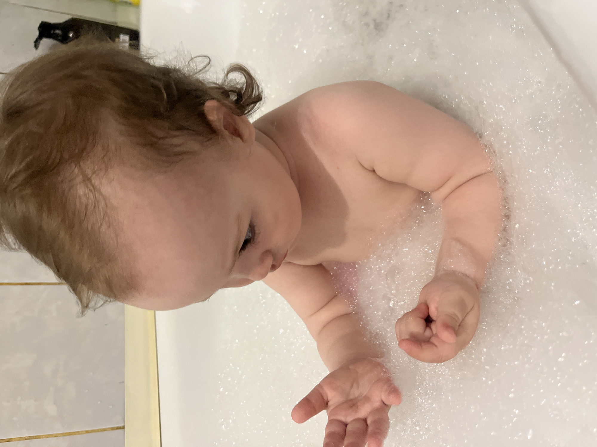 A baby is in a bathtub filled with bubbles. The baby has light skin and brown hair. The baby is leaning forward with their arms extended, and one hand is open while the other is closed in a fist. The location appears to be a private residence and not a public place in Shushary, Russia. The environment is simple and focused on bathing.