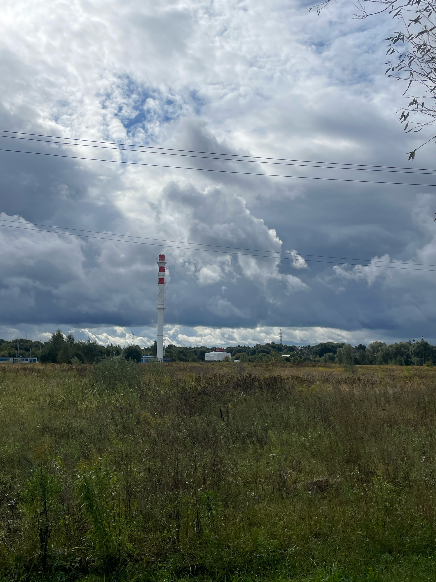 The image depicts an outdoor scene with a tall, white chimney with red bands, standing in a field of tall grass and vegetation. The sky is filled with dramatic, dark clouds, suggesting overcast or stormy weather. In the background, a line of trees and some buildings are visible. Power lines stretch across the upper portion of the image. The overall impression is one of an industrial or semi-industrial area on the outskirts of a settlement, with a focus on natural elements like the field and the sky. The location is identified as Fabriki 1 maya, Russia.