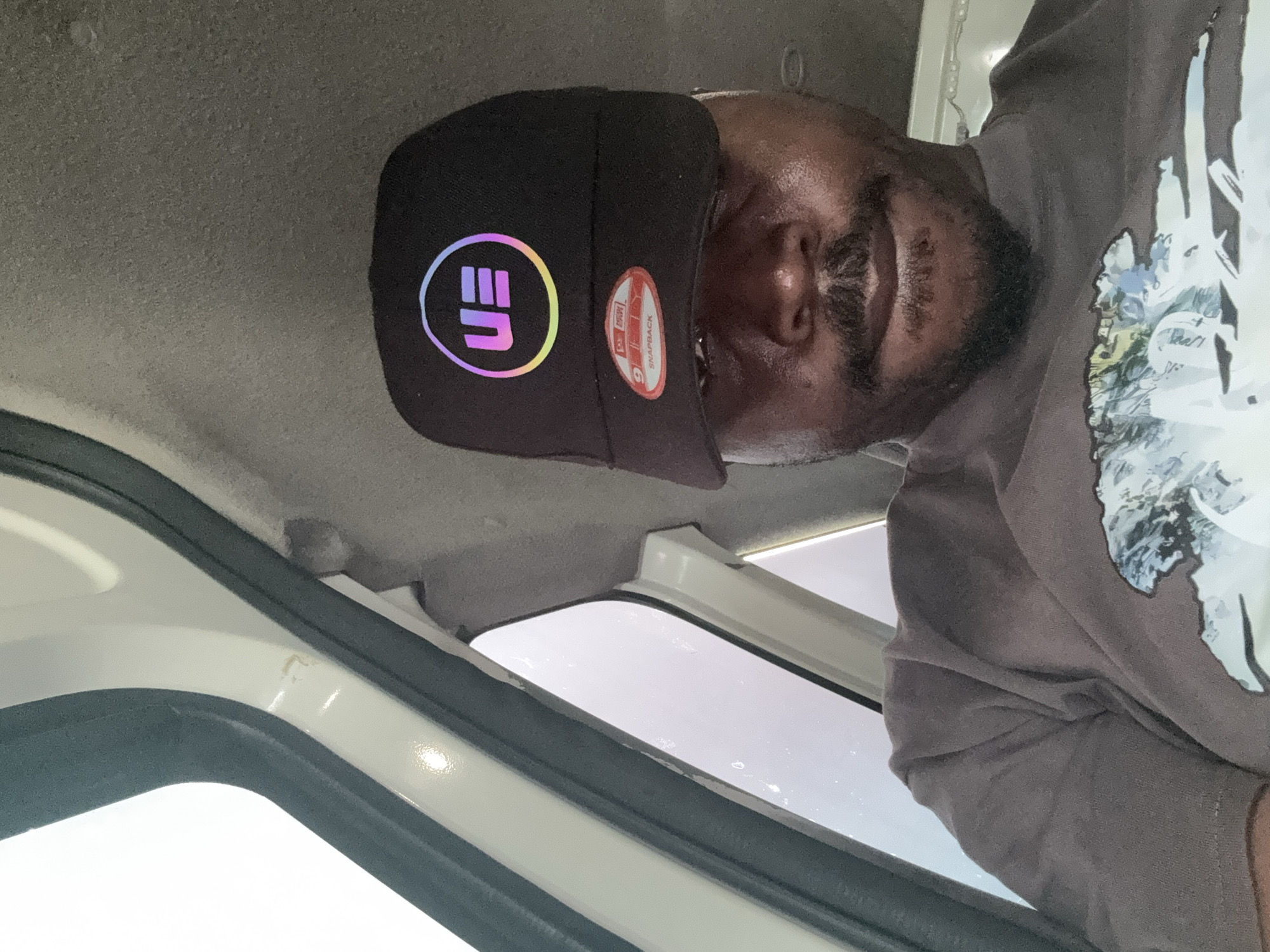 A medium, high-angle shot, taken inside a light-colored vehicle, with the camera pointed towards a dark-skinned man wearing a black baseball cap and a brown long-sleeved shirt with a white graphic design. The man is facing the camera with his head tilted. The black baseball cap has a distinctive circular emblem with the letters "EU" in a colorful gradient. The interior of the vehicle shows a window frame. The image does not provide visual confirmation of the location in Ijanikin, Nigeria.