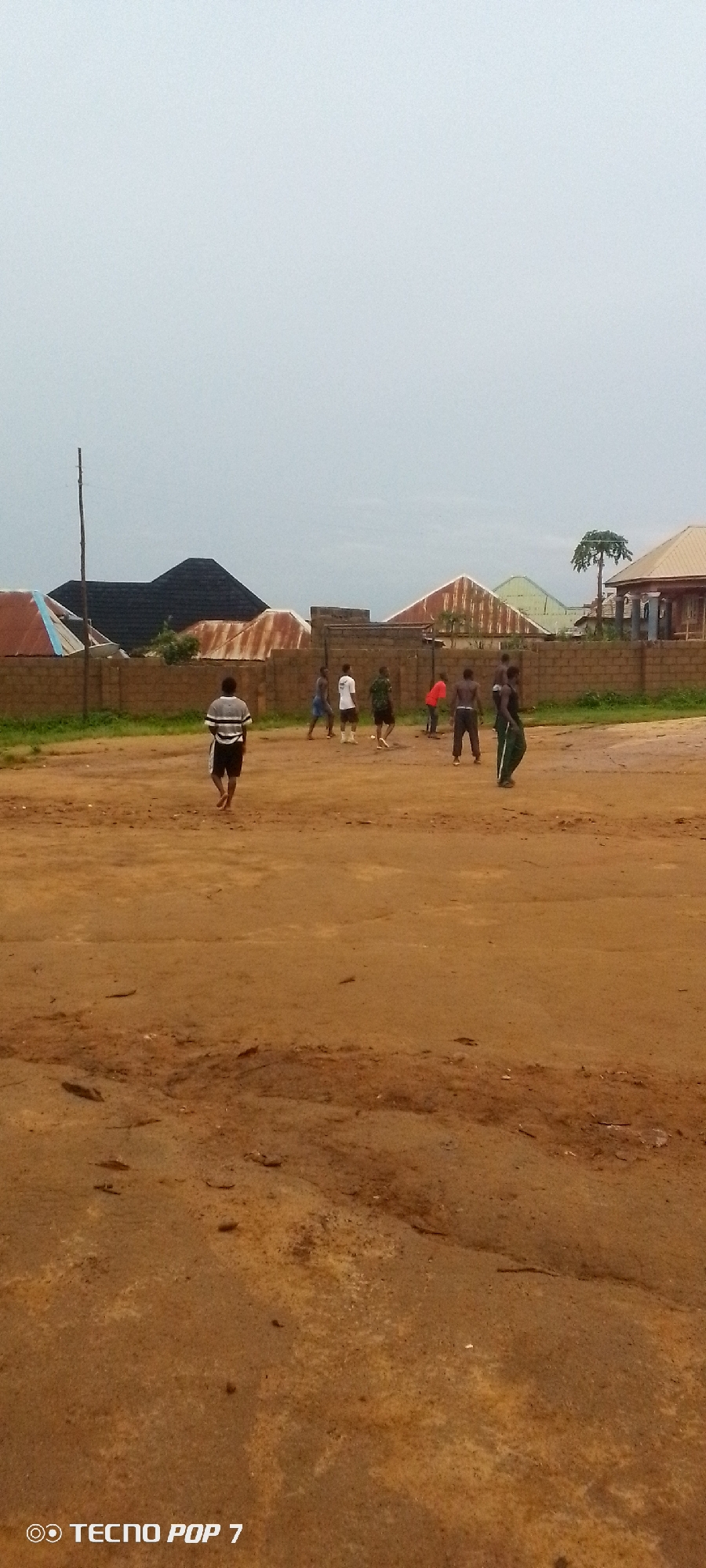In an outdoor scene in Unknown, Unknown, several young males are playing a game of soccer on a dirt field. A tall, thin pole stands to the left of the frame. In the background, various houses with corrugated metal roofs are visible, separated by a brick wall. The sky is overcast and pale. The foreground is a dusty, uneven dirt surface.