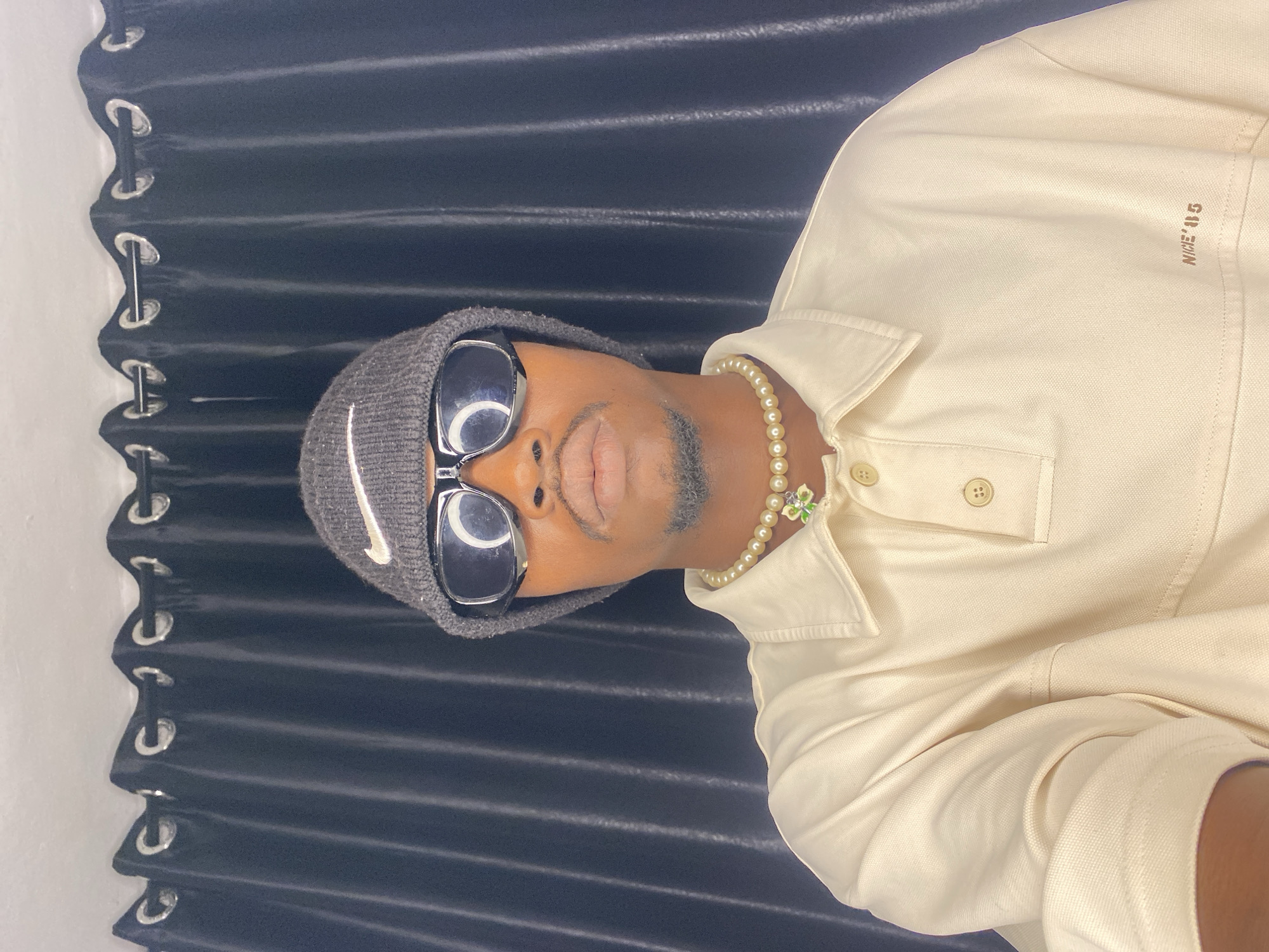 In Abakaliki, Nigeria, a young Black man with a goatee is photographed from the chest up. He is wearing a grey Nike beanie, black wrap-around sunglasses, and a cream-colored collared shirt with two buttons at the neck. Around his neck, he wears a pearl necklace with a green clover pendant. The background is a dark, pleated curtain. The lighting is bright, creating reflections on his sunglasses.