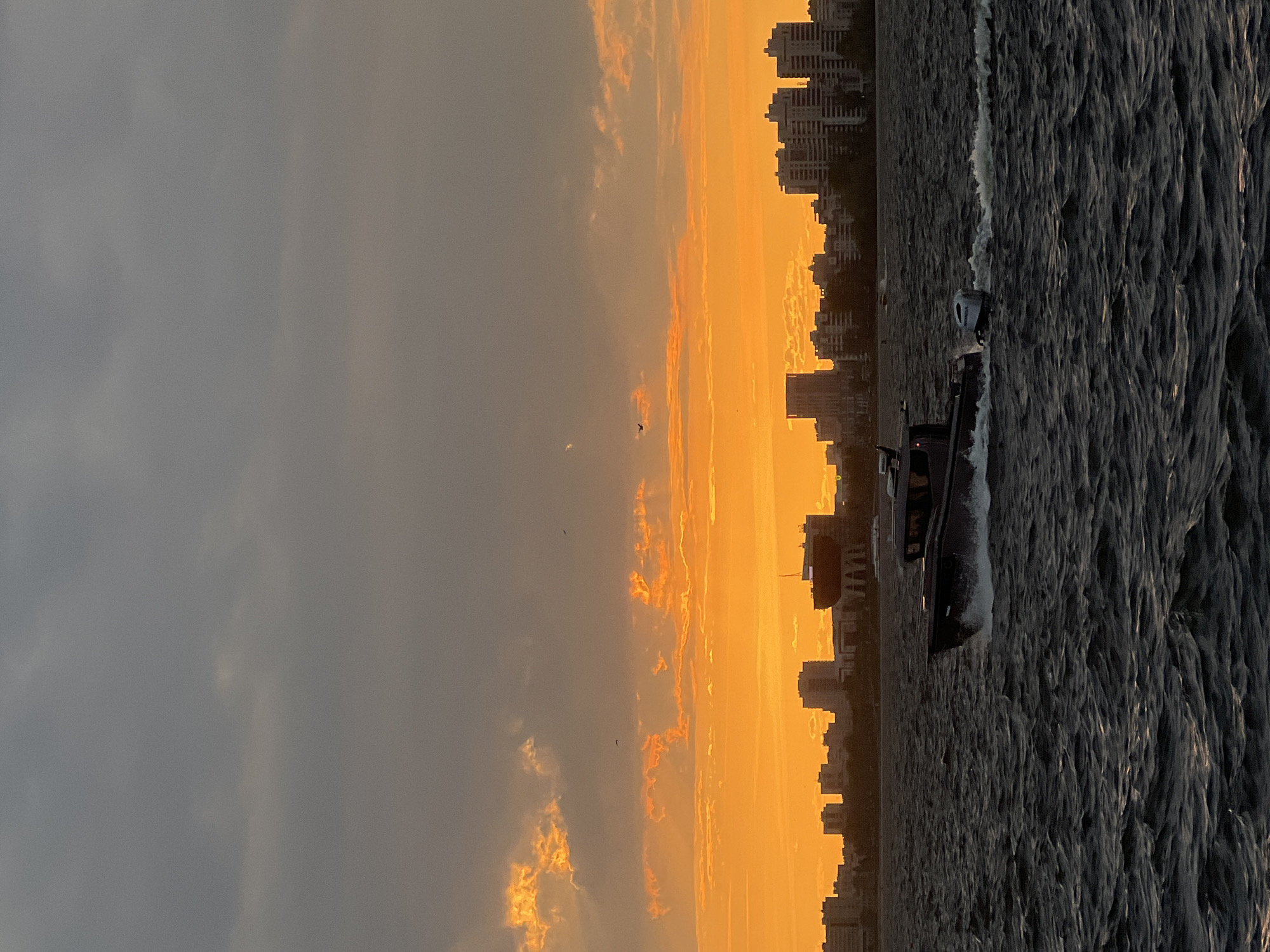The image shows a body of water with a speedboat moving across it, leaving a wake. On the opposite shore, a cityscape silhouette is visible against a golden, cloudy sky. The overall scene suggests a sunset or sunrise setting. It is difficult to conclusively place the scene in Kazan, Russia, without additional evidence from the image. No people are visible in the image.