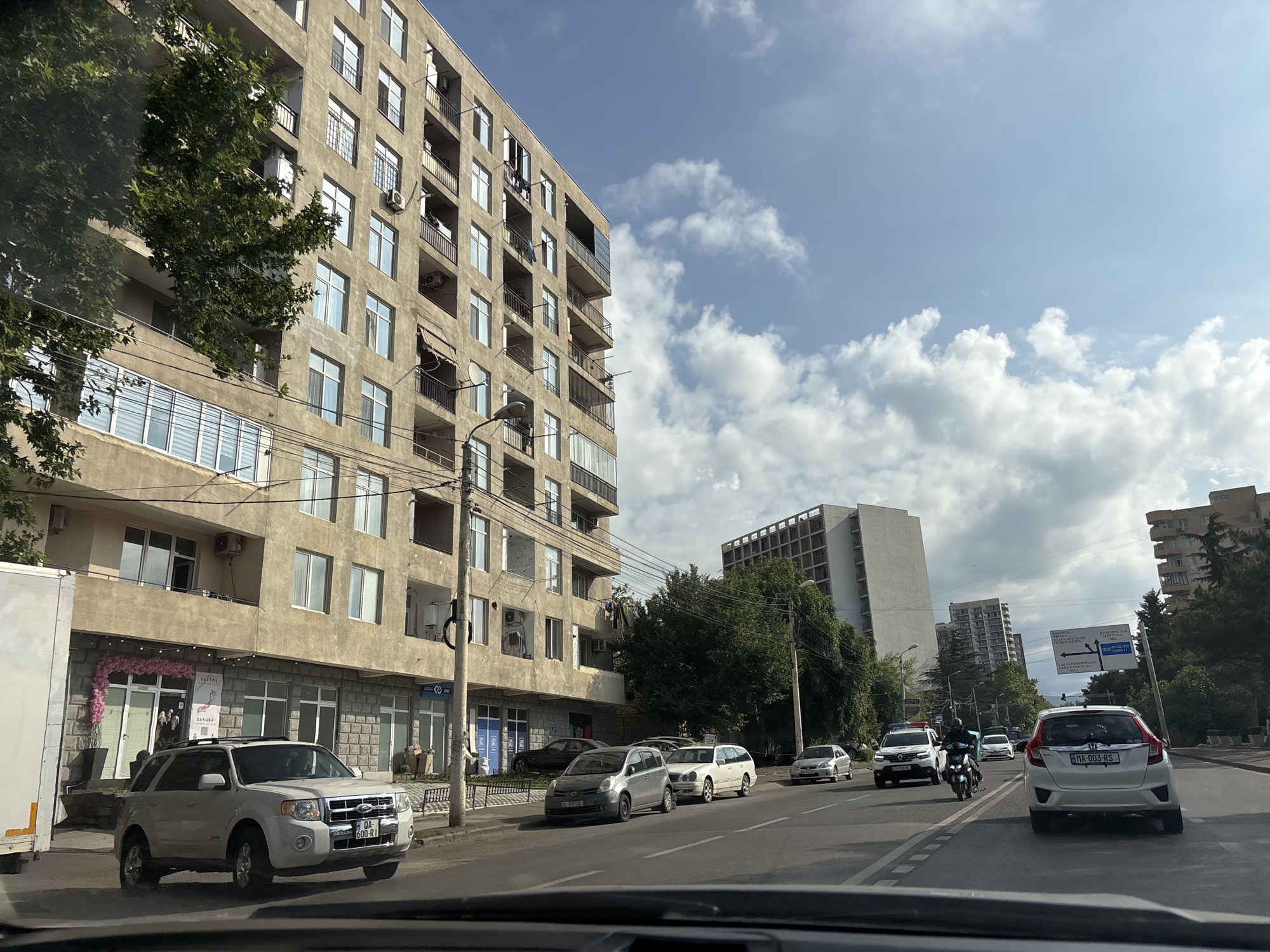 The image shows a street scene in Tbilisi, Georgia. The street is lined with multi-story buildings on the left, with some trees and other buildings in the background. Several cars are visible on the road, including a white SUV in the foreground, a white van, a silver car, and a white hatchback. A motorcycle with a rider wearing a helmet is also present. The sky is partly cloudy. The buildings appear to be residential or mixed-use, with storefronts on the ground floor. The overall impression is that of a typical urban environment.