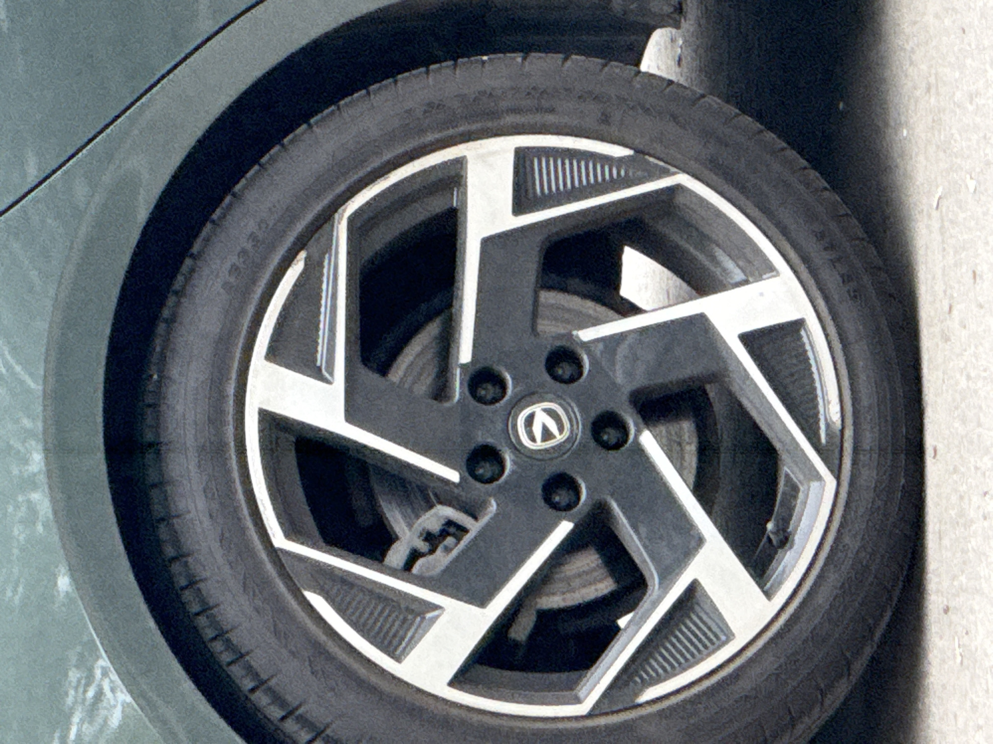 The image shows a close-up of the front wheel of a vehicle. The wheel has a five-spoke design in black and silver, with a prominent car brand logo at the center. The tire appears to be black. The wheel is situated within the car's wheel well, with a portion of the vehicle's body visible. The image is taken in outdoor lighting. It is unlikely that this image is from Moscow, Russia, unless further information is provided to support that claim.
