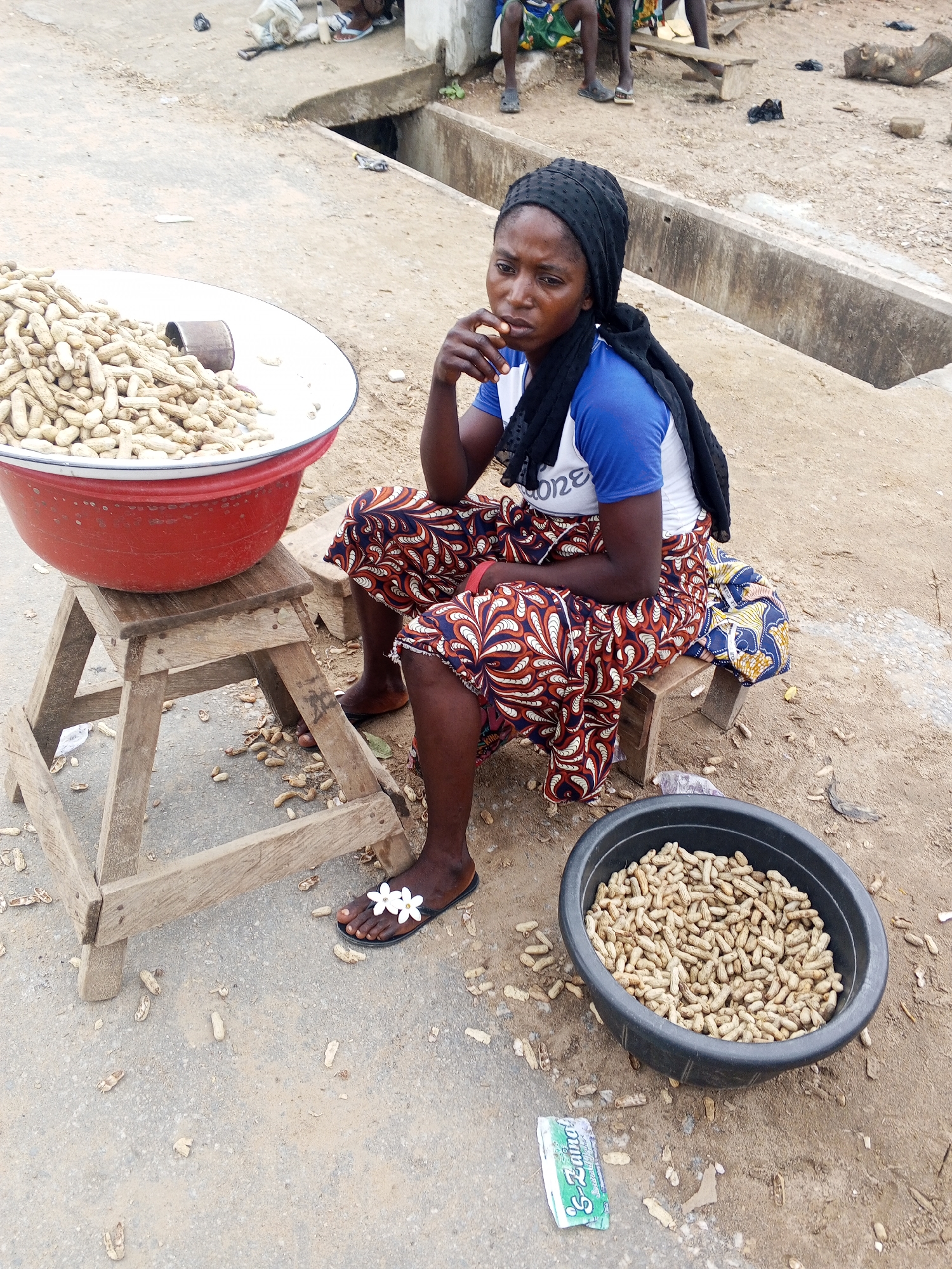In Zango-ndaji, Nigeria, a Black woman sits on a wooden stool beside a large red basin filled with peanuts. She wears a black headscarf, a blue and white t-shirt, and a patterned skirt. Her feet are adorned with flip-flops featuring white flower embellishments. Scattered peanuts and shells litter the sandy ground around her. To her right, a black oval basin also holds a quantity of peanuts. In the background, other individuals are visible, and concrete structures line the area. The overall scene depicts a street-side vendor preparing to sell peanuts in a bustling environment.