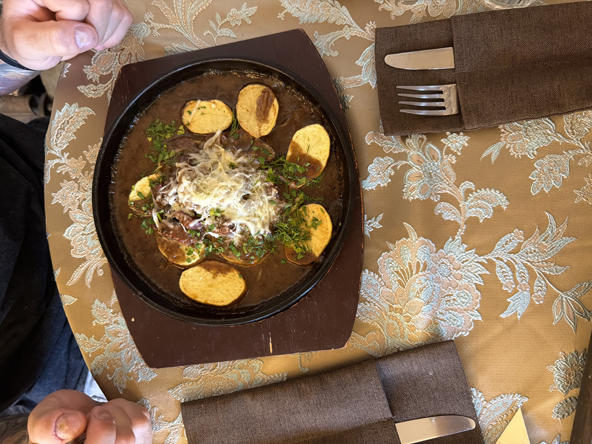 A close-up, overhead shot shows a sizzling platter of meat and potatoes on a table with a floral patterned tablecloth. The platter, a dark, round, cast-iron dish with a wooden base, contains a rich brown sauce with pieces of meat, topped with shredded cheese and chopped green herbs. Sliced, golden-brown potatoes are arranged around the edge of the platter. To the left, a brown napkin is folded with a single silver knife. To the right, another brown napkin holds a silver fork and knife set. In the background, parts of two people are visible; one person's hands are in the lower left of the frame, appearing to hold cutlery, and the other person's hands are in the upper right, with visible tattoos. The table is set for a meal. The scene appears to be in a restaurant in Suzdal, Russia, a city known for its historic architecture and traditional cuisine.