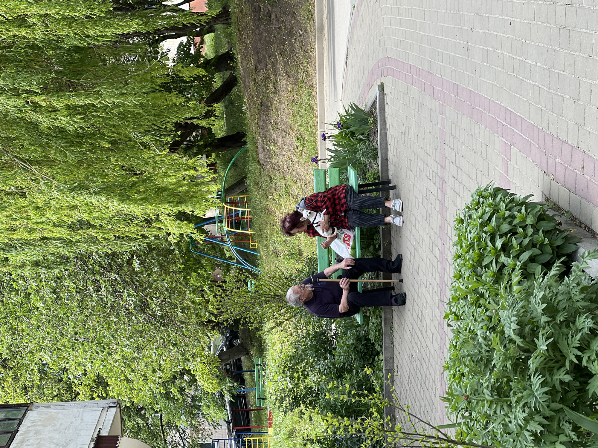 Two elderly individuals are sitting on a green bench in a public space. One individual is holding a small white dog. The other individual is holding a wooden cane. Behind the bench is a playground with colorful equipment. Brick pavers compose the walking path in the park. The location is Ternopil, Ukraine.