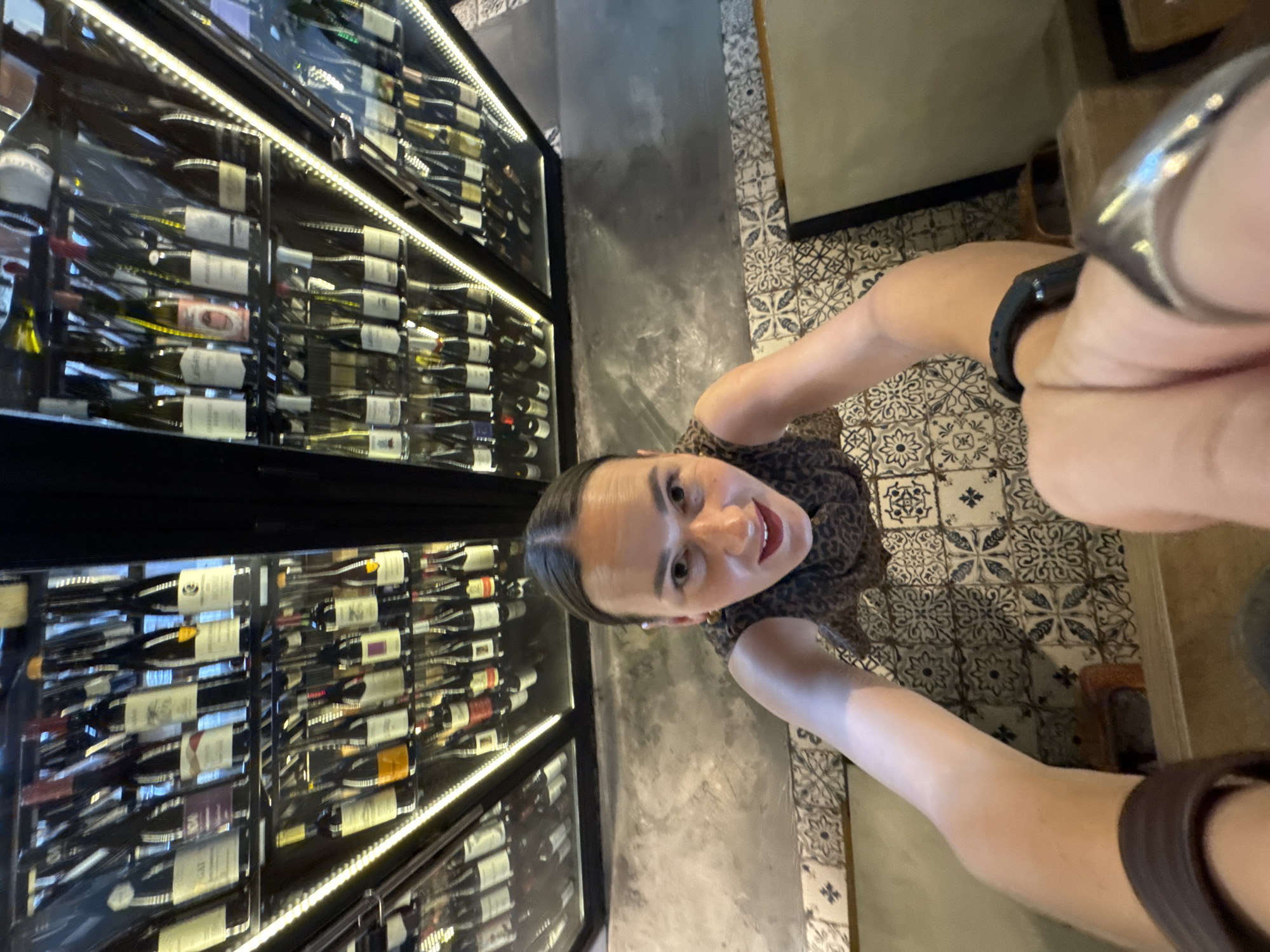 A woman, in a close-up, from a high angle perspective, is captured in an image taken from within a wine cellar. She is smiling at the camera, her head tilted slightly. She is wearing a patterned top and a watch on her left wrist, and a ring on her right index finger. The background consists of rows of wine bottles neatly arranged on shelves, illuminated by bright interior lighting. The floor is a mosaic tile pattern, and there are glimpses of concrete surfaces. The image is an indoor scene, likely a restaurant or a wine storage facility. The city of Sirius, Russia, is not depicted or referenced in the image.
