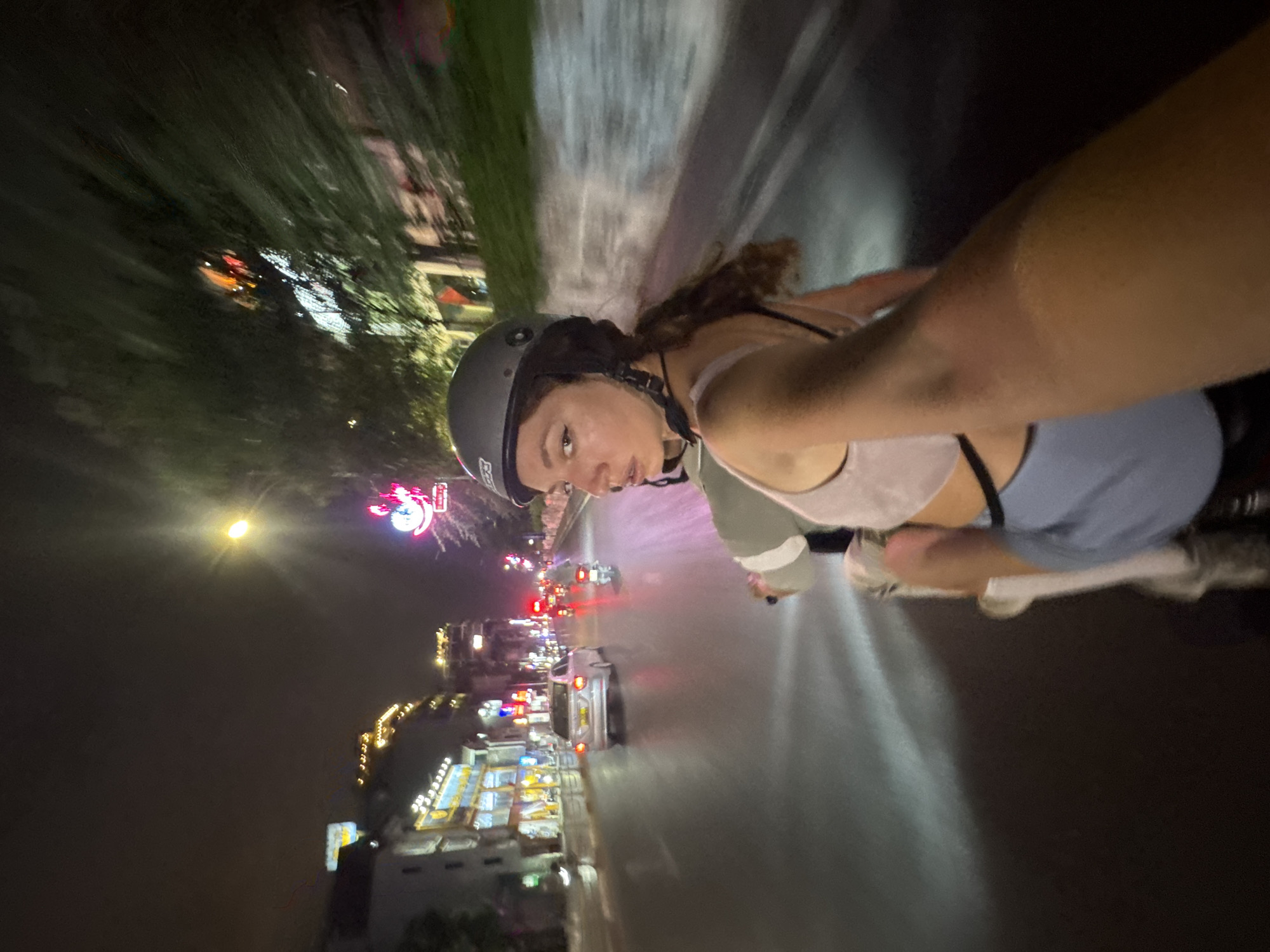 The image depicts a woman riding on the back of a motorcycle at night in Hanoi, Vietnam. She is wearing a gray helmet, a light pink crop top, and blue shorts. Her body is angled away from the camera, and she is looking back over her shoulder towards the viewer with a neutral expression. The motorcycle appears to be in motion, as evidenced by the motion blur in the background.
The setting is a city street illuminated by streetlights and neon signs. Buildings line the left side of the street, with some displaying illuminated windows and signage. Several other motorcycles and a silver car are visible on the road, suggesting moderate traffic. The road surface appears wet, reflecting the lights. The general atmosphere is that of a bustling urban environment at night.