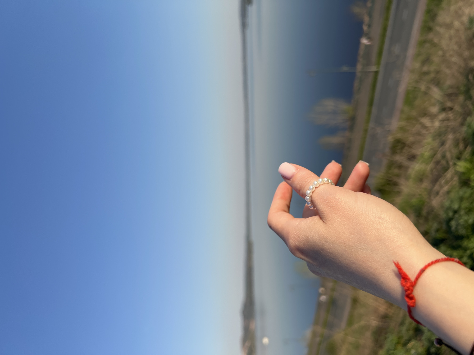 A hand wearing a pearl ring and a red knotted bracelet is held up against a backdrop of water, sky, a road, and grassy land. The location is Sviyazhsk, Russia.