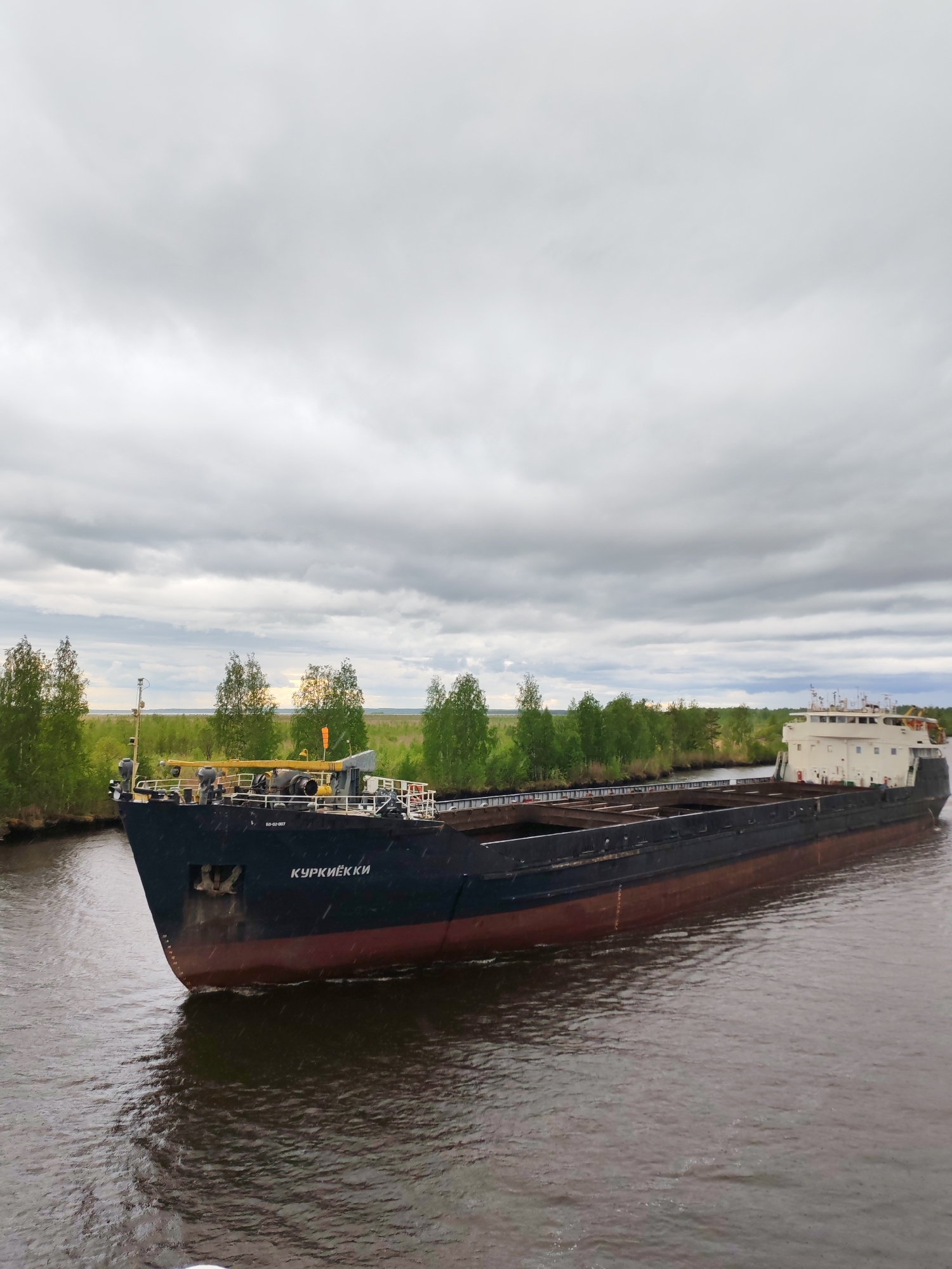 A barge, identified with the name "КУРКИЁККИ" painted in white on its bow, is moving through a waterway in what appears to be Ozernoe Ust'e, Russia, given the style of lettering on the ship. The vessel is long, black and red, and appears to be empty. The water is dark and choppy. The barge is navigating past a bank lined with green trees and foliage. Overhead, the sky is overcast with gray clouds.