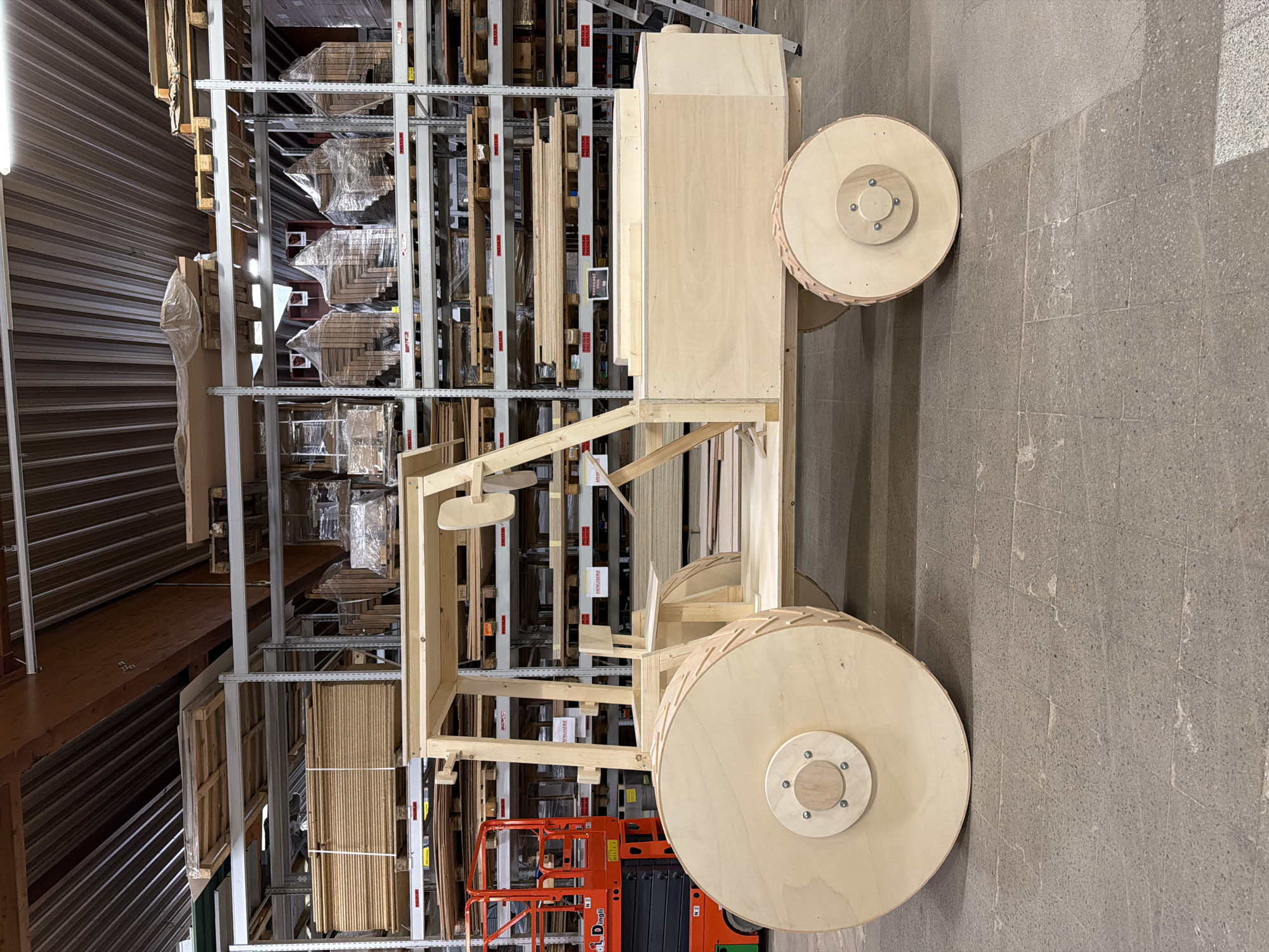 The image shows a large, unfinished wooden tractor model in a warehouse or lumberyard. The tractor is constructed from plywood and dimensional lumber, with large wooden wheels. The interior of the tractor appears to be hollow, with a simple wooden seat. In the background, tall industrial shelving units are filled with stacks of wood products, including plywood sheets and other lumber. To the left of the tractor, a red scissor lift is partially visible. The flooring is made of grey concrete tiles. The overall scene suggests a workshop or storage facility for building materials, potentially in the city of Leudelange, Luxembourg, given the context of the provided information. There are no people or animals visible in the image. The lighting is industrial, consistent with a warehouse environment. Labels are visible on some of the shelves, though the text is not clearly legible.