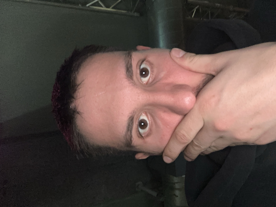 An individual, identified as male, is depicted in a tight, head-and-shoulders crop. He has fair skin, wide brown eyes with dilated pupils, and short dark hair with visible reddish-purple highlights. Light stubble is present on his chin and upper lip. His right hand is positioned over his mouth, covering the lower face, with fingers pressed against the lips and the thumb resting on his left cheek/jawline. He is wearing a dark, possibly black, hooded garment, visible around his neck and left shoulder. The background is dimly lit and indistinct, revealing industrial or utility elements. On the left, a light-colored pipe or conduit is partially visible. On the right, a metallic structural lattice, possibly part of a truss or scaffolding, is discernible in the upper corner, along with a horizontal pipe behind the subject's right shoulder. This scene is located in an unspecified indoor environment within the city of Varna, Bulgaria.