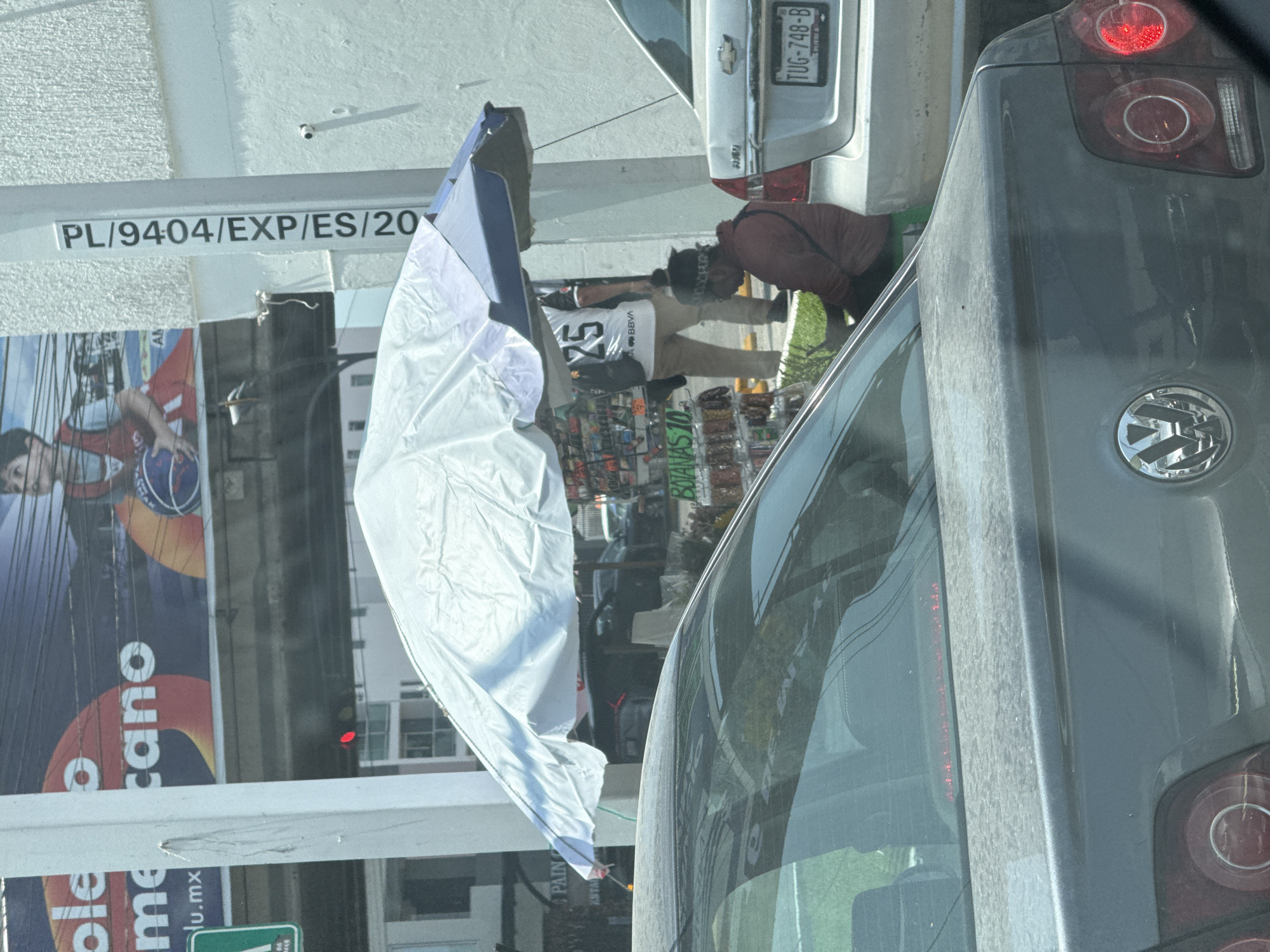 The image depicts a street scene likely in Puebla, Mexico, based on the context of the provided information. A grey Volkswagen car, partially visible, is positioned on the right side of the frame. Next to it, partially obscured, is a white Chevrolet car with license plate TUG-748-B. A street vendor is visible near the Chevrolet, wearing a jersey with the number "25" and the BBVA logo, attending to a makeshift stall that features a white canopy. The stall appears to be selling various items, including snacks or magazines. A building is present in the background, displaying the text "PL/9404/EXP/ES/20" and parts of the word "ole" and the phrase "me cano lu.mx" from an advertisement.