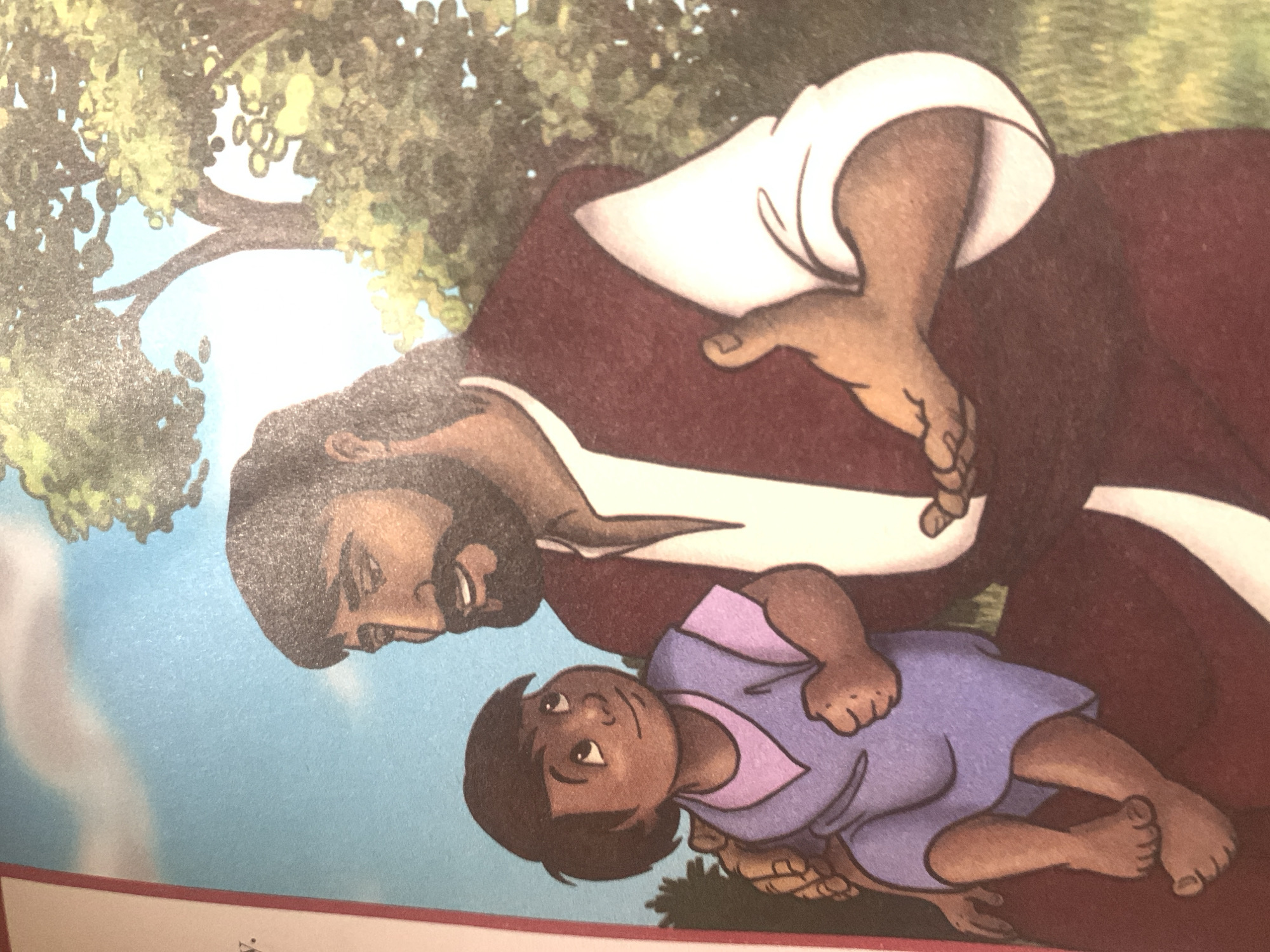 The image depicts an illustration of Jesus Christ interacting with a young child outdoors. Jesus, with a beard and long hair, is wearing a maroon robe over a white garment. He is seated and holding the child on his lap. The child, with dark hair and brown skin, is wearing a lavender tunic with a pink undershirt and is looking up at Jesus. Jesus's right hand is extended towards the child, palm open, in a gesture that suggests teaching or comfort. The background shows a blue sky, green trees, and a patch of grass. The style is illustrative, suggesting it is from a book or religious material. There is no indication of the city of Jauro Yinu, Nigeria, within the image.