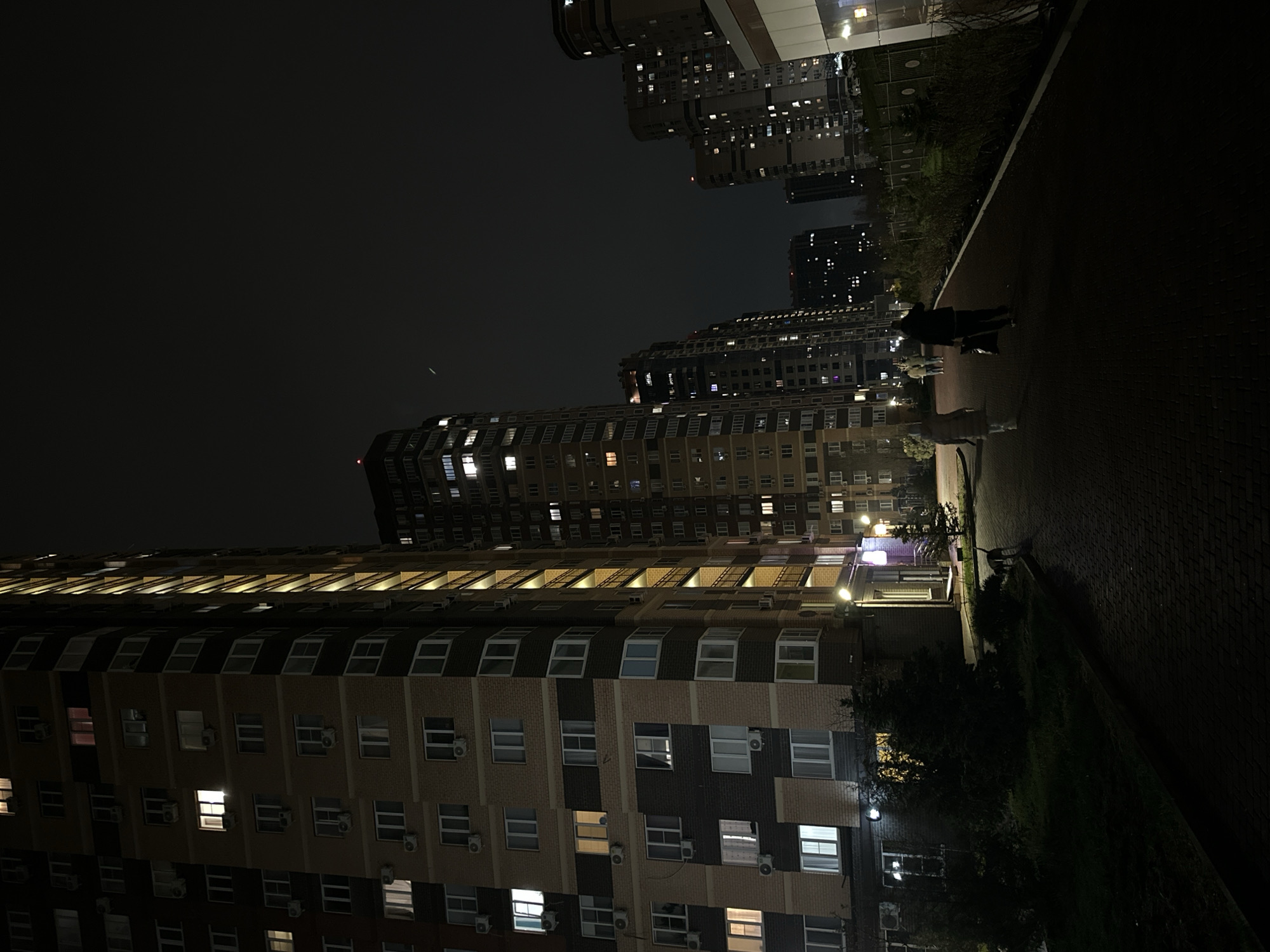 The image shows a nighttime view of a residential area in Rostov-on-Don, Russia. Several high-rise apartment buildings are visible, many with illuminated windows. A paved walkway runs along the right side of the image, with a dark, silhouetted figure walking on it. Some greenery, including trees and bushes, is present along the walkway. The sky is dark. Air conditioning units are fixed on building walls.
