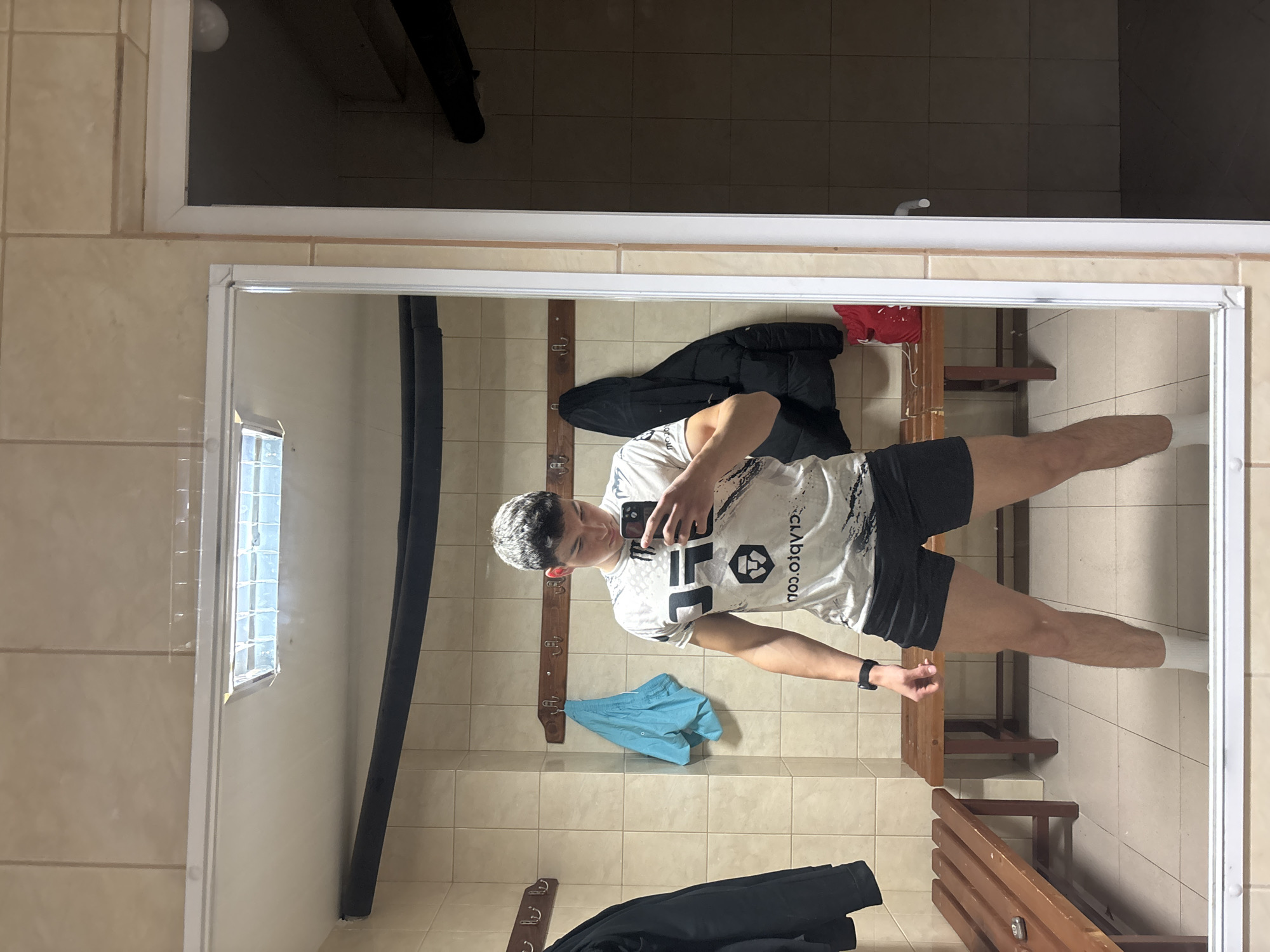 A young male is captured in a mirror selfie within what appears to be a locker room or changing facility in Zlatograd, Bulgaria. The subject, seen from the waist up in the mirror's reflection, stands centrally, holding a smartphone to take the picture. He has short, dark hair with lighter highlights and is wearing a white and black graphic t-shirt, black shorts, and white socks. A dark smartwatch is visible on his left wrist.
The setting features light beige square tiles on both the walls and floor. Wooden benches and wooden coat racks line the walls. A black jacket is draped over a bench in the foreground, and a light blue garment, possibly shorts or a towel, hangs on a hook behind the subject. A red item is also partially visible on another bench. To the left, a narrow window with glass block panes provides some natural light. A black insulated pipe runs vertically along one wall. The ceiling area reflected above the mirror is dark, with a visible light bulb.
The t-shirt features a "UFC" logo and the mirrored text "CLABRO.COM". The scene suggests a functional, well-lit indoor environment, characteristic of a sports or gym changing area.