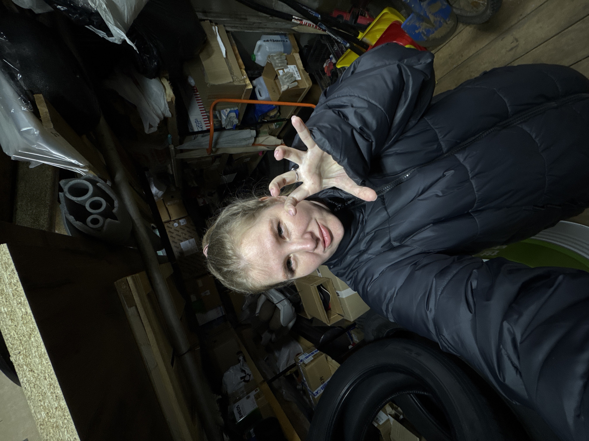 A person with fair skin, blonde hair pulled back, and wearing a dark puffer jacket is holding their hand up with fingers spread. They appear to be in a cluttered storage area or garage. Various cardboard boxes, containers, and other miscellaneous items are visible in the background. A tire lies near the person. The location is Gornyi (Dvurechenskoe S/P), Russia.