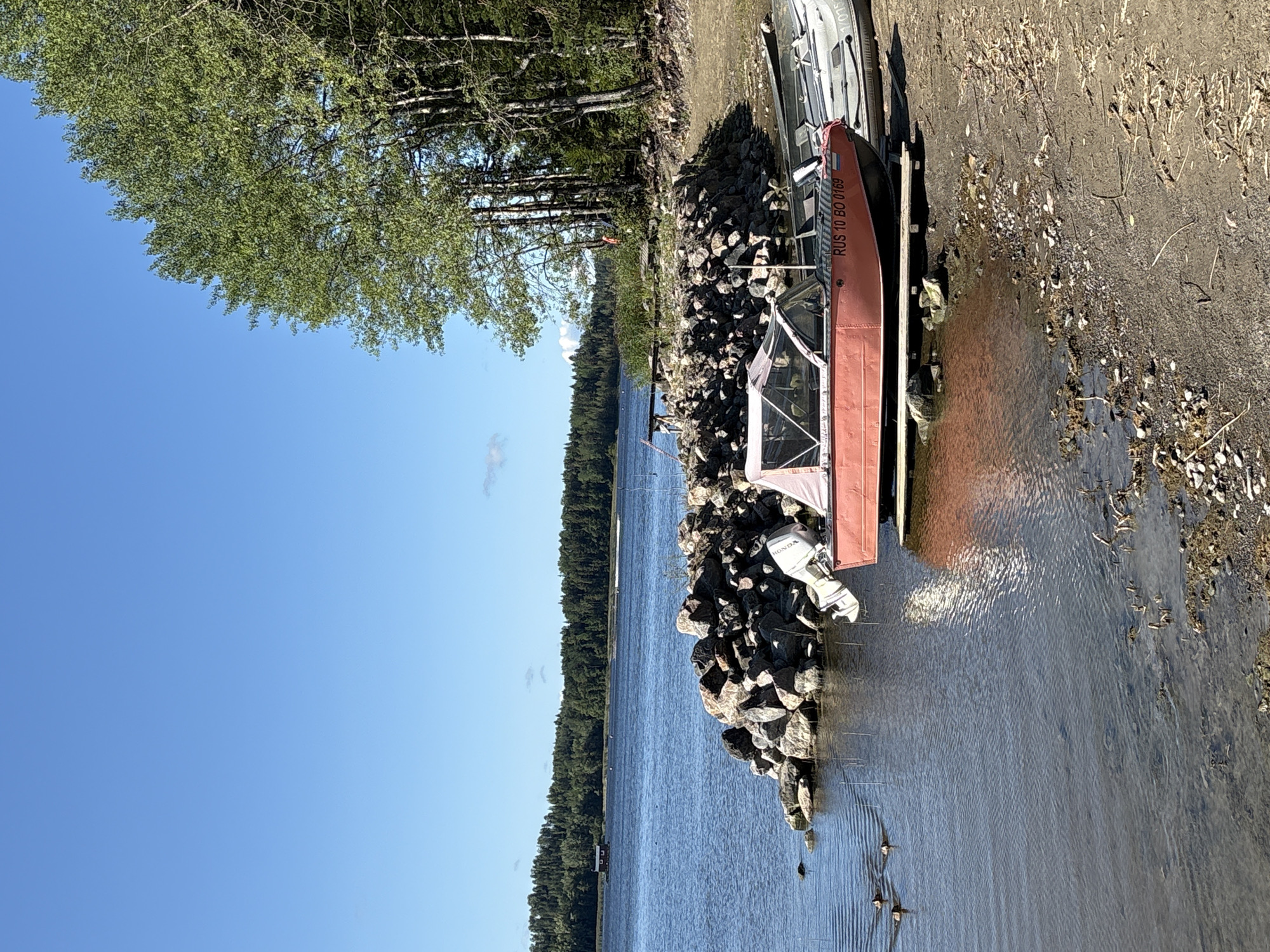 The image depicts a sunny day on the shore of a lake in Rautalakhti, Russia. The foreground is a rocky beach with a shallow waterline. A red motorboat with a white canopy is partially submerged and resting on wooden planks near the shore. Another silver boat is docked behind it. In the water, a small group of ducks is swimming. The middle ground features a rocky embankment leading to a wooden structure with a walkway. The background shows a dense forest on the opposite shore, with a small red building visible on the water in the distance. The sky is a clear, bright blue. The overall scene is peaceful and evokes a sense of a quiet summer day by the water.