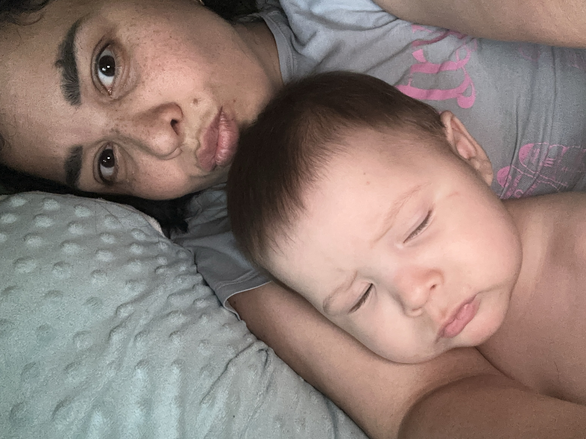 A selfie-style photograph depicts a woman and a baby lying down on what appears to be a bed covered with a light-colored textured blanket. The woman, positioned behind the baby, has brown hair and freckles on her face, with one eye open and looking directly at the camera. Her lips are pursed. The baby, positioned in front of the woman, is asleep with its eyes closed, lying on its side with its head resting on what appears to be the woman's arm. The baby has dark hair and fair skin. Both the woman and the baby are wearing casual clothing. The environment suggests a domestic setting.