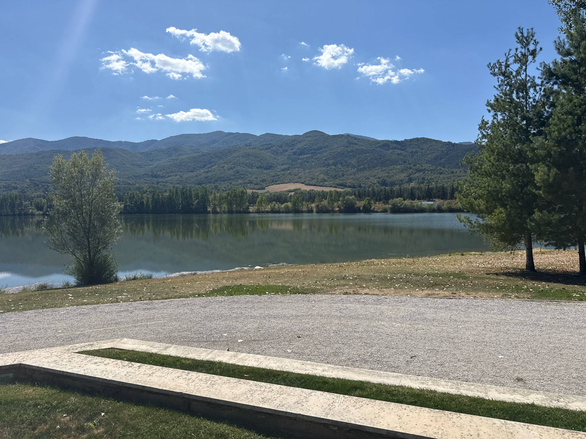 A serene landscape view depicts the lake in the city of Pravets, Bulgaria, under a bright, partly cloudy sky. In the foreground, a concrete structure with a grassy area is visible, leading to a gravel path. Beyond the path lies the tranquil surface of the lake, reflecting the surrounding trees and the distant, verdant mountains. A lone tree stands to the left on the lake's edge, while two pine trees are prominent on the right side of the image. The scene is characteristic of a peaceful natural setting, likely a park or recreational area.