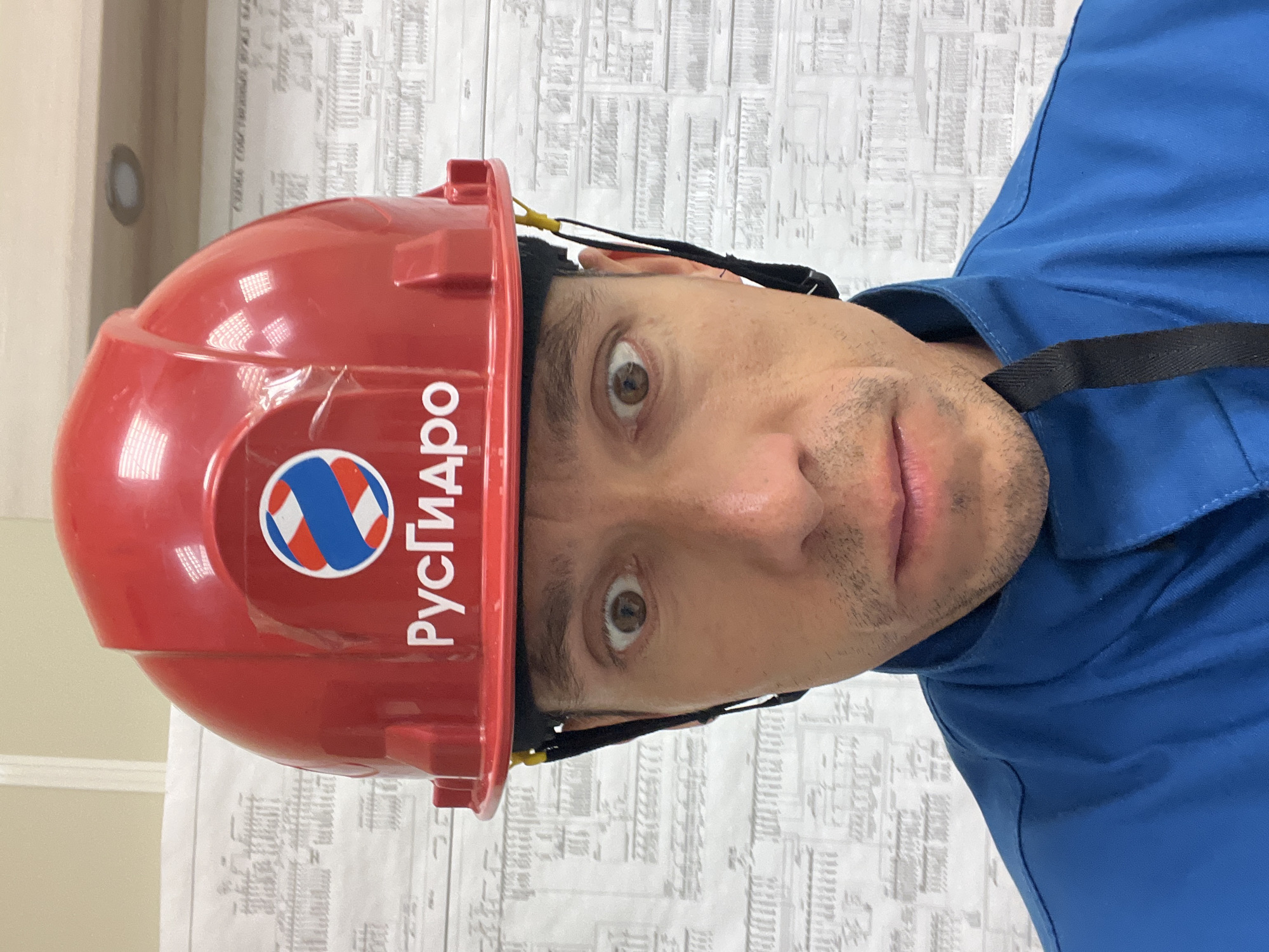 A man wearing a red hard hat with the "RusHydro" logo and a blue uniform is looking directly at the camera. The hard hat has a circular logo with red, white, and blue diagonal stripes. The background is a wall covered with technical blueprints. The man has wide eyes and is looking upwards and slightly to his left. The image appears to be taken indoors. The city of Chaikovsky is known for its industrial sector, including hydroelectric power plants, which aligns with the "RusHydro" branding visible on the hard hat.