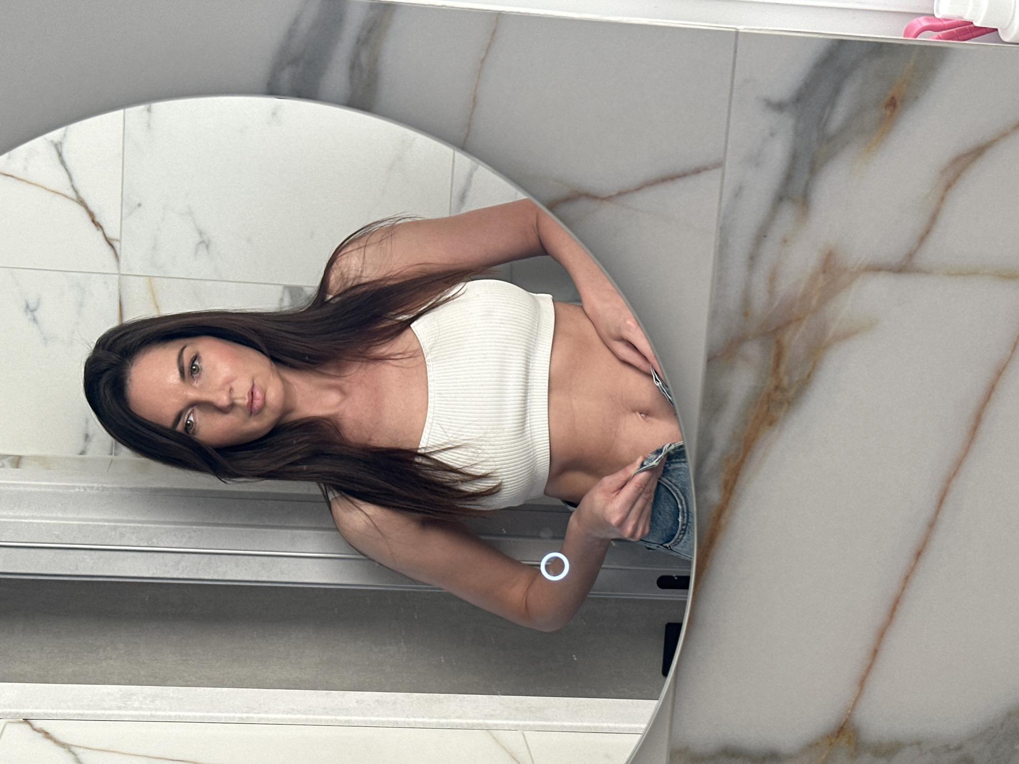 A woman with long brown hair, wearing a white ribbed tank top and blue jeans, is reflected in a round mirror. The background consists of white marble tiles with brown and gray veining. A shower door or glass panel is partially visible in the reflection. The location is a bathroom. The image does not provide enough information to determine if it is specifically in the city of Chaikovsky, Russia.