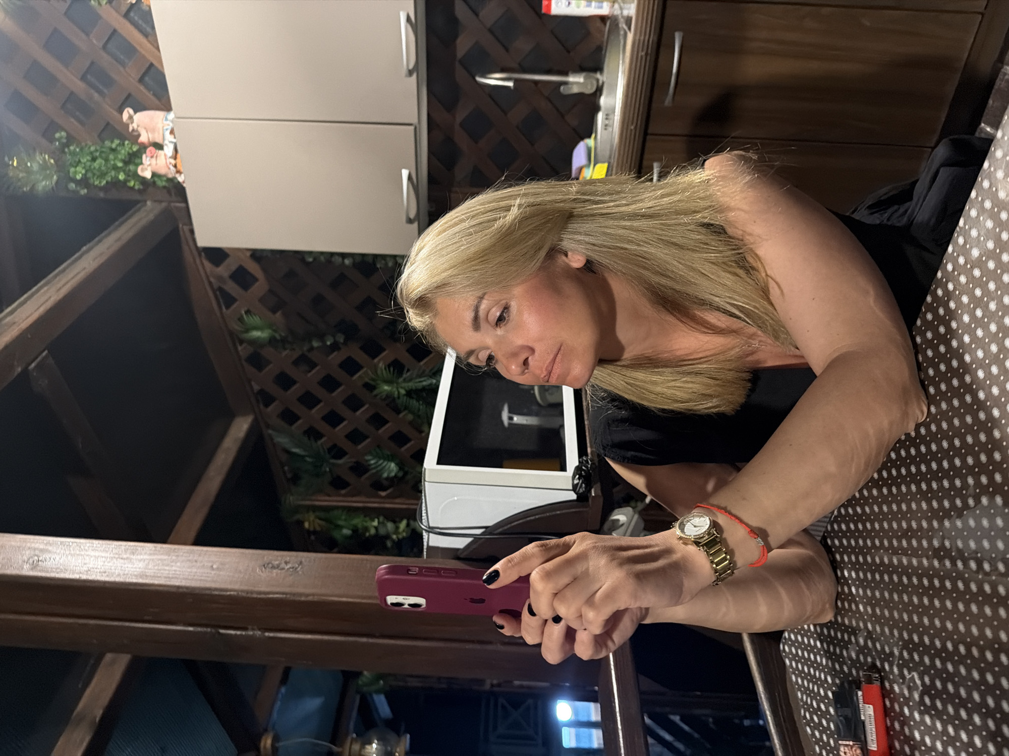 A woman with blonde hair is sitting at a table, holding a smartphone in her hands. She is wearing a black dress and a gold watch on her left wrist. A red string bracelet is also visible on her left wrist. The table has a patterned surface. In the background, there is a kitchen area with cabinets, a sink, and a microwave. There are also decorative pig figurines on top of the cabinets. The scene appears to be on an outdoor patio or balcony. The presence of a lighter and cigarette pack on the table suggests a possible smoking area. The environment suggests a relaxed, informal setting, possibly a private residence or a cafe with an outdoor seating area. The lighting indicates it is either evening or the area is dimly lit.