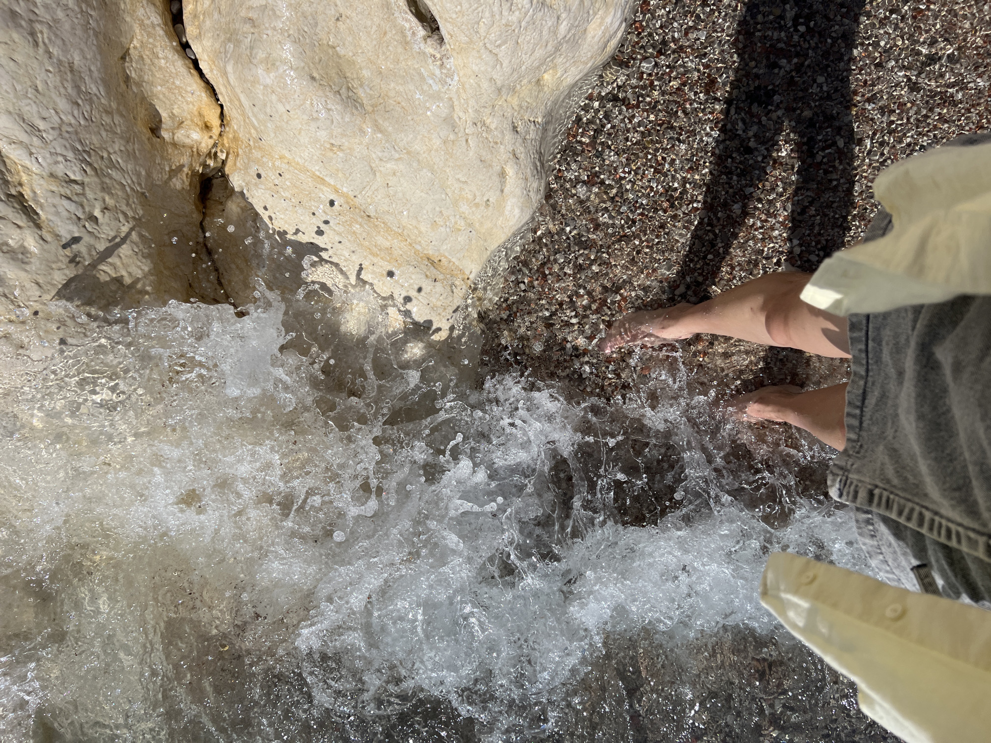 A first-person, high-angle shot captures a person's bare legs and feet partially submerged in shallow, clear water at a pebble beach. The person is wearing light-colored denim shorts or a skirt and a pale yellow top or garment draped over their lap. The water in the foreground and center is actively splashing and foaming, indicating a wave or current. To the right, large, light-colored limestone rocks with visible cracks and dark stains form a portion of the shoreline. The pebbles on the beach are small and varied in color. The scene is brightly lit by natural light, casting shadows from the rocks onto the water and pebbles. The setting is Mirište, Montenegro.
