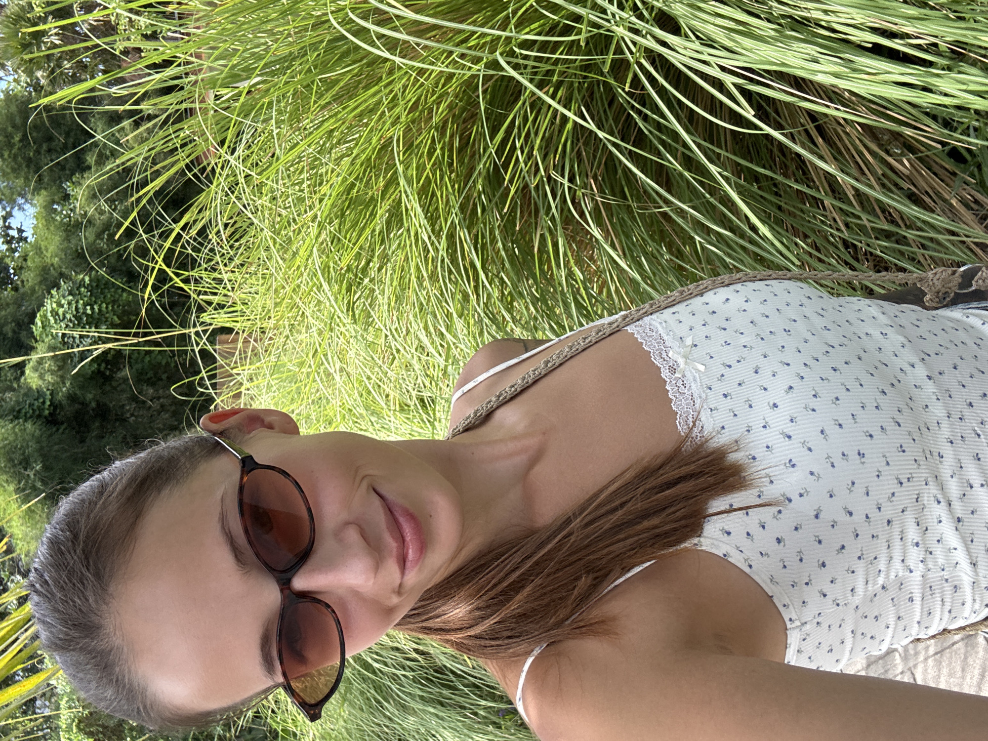 A young woman with fair skin and brown hair is depicted in a medium shot, lying on her side in a grassy area. She wears sunglasses with brown frames and a white, floral-patterned top with lace trim. A woven shoulder strap is visible across her chest. The background consists of tall, green grass and foliage suggesting a garden or park setting. The location is inferred to be in the city of Mengwi, Indonesia, though specific geographical features are not discernible within the frame.