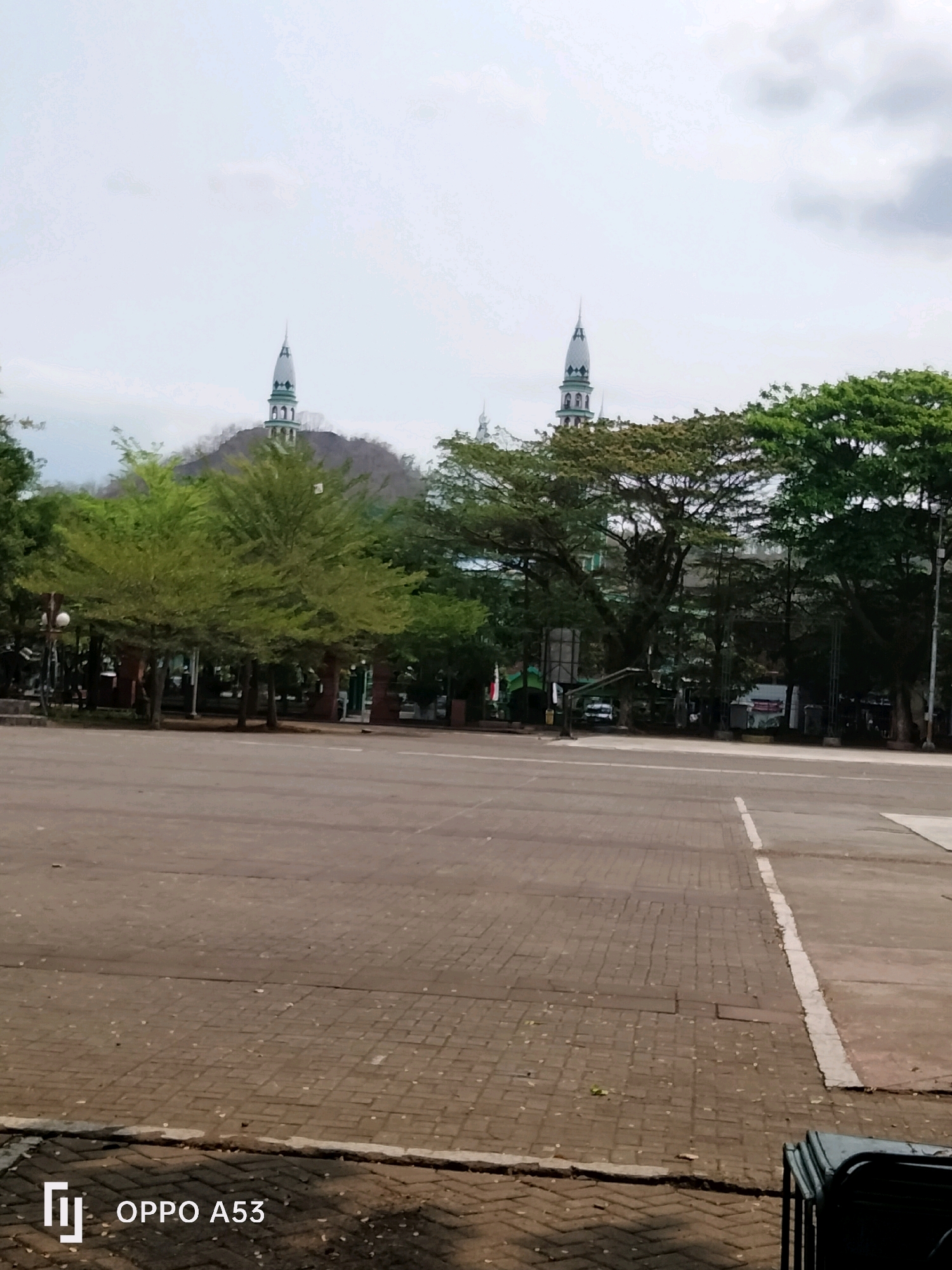 The image shows a wide paved plaza with a tree-lined background featuring two mosque minarets. The plaza is paved with interlocking bricks, and there are some scattered leaves. In the background, mature trees with dense green foliage are visible, some obscuring the lower portions of buildings. Behind the trees, two tall, slender mosque minarets with white and green accents rise into a pale, overcast sky. The minarets are situated behind a hilly, tree-covered incline. The environment appears to be a public space, possibly adjacent to a religious or civic building complex. The bottom right corner of the image shows a stack of dark chairs. The lower left corner displays the "OPPO A53" watermark. The scene is devoid of people. This image was captured in Trenggalek, Indonesia.