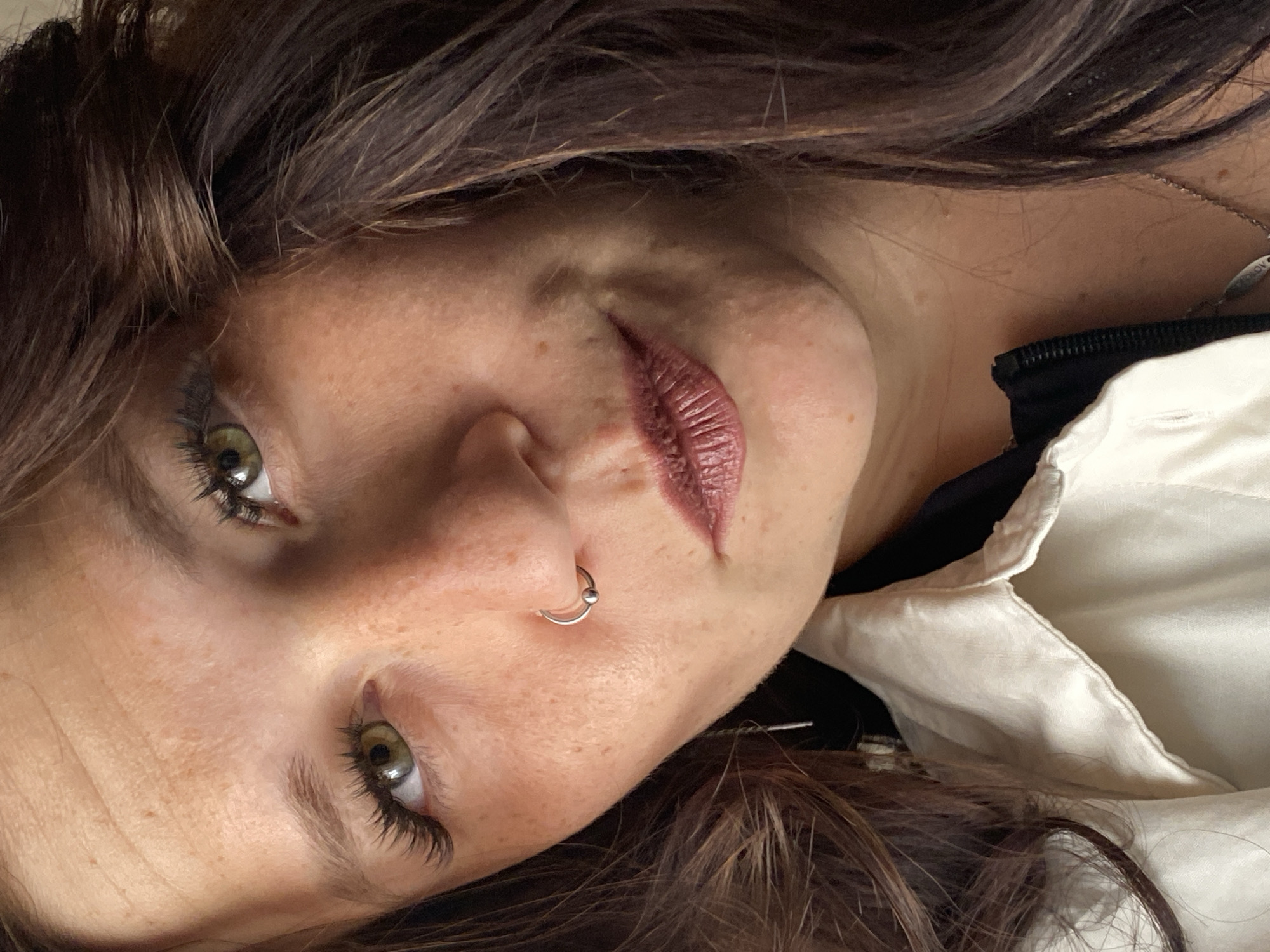 The image is a close-up portrait of a woman with brown hair and green eyes. She is wearing a nose ring and has visible freckles on her face. She is also wearing a light-colored collared shirt over a dark jacket. The background is blurred and does not provide specific environmental details. The image does not contain any information about the city of Chaikovsky, Russia.