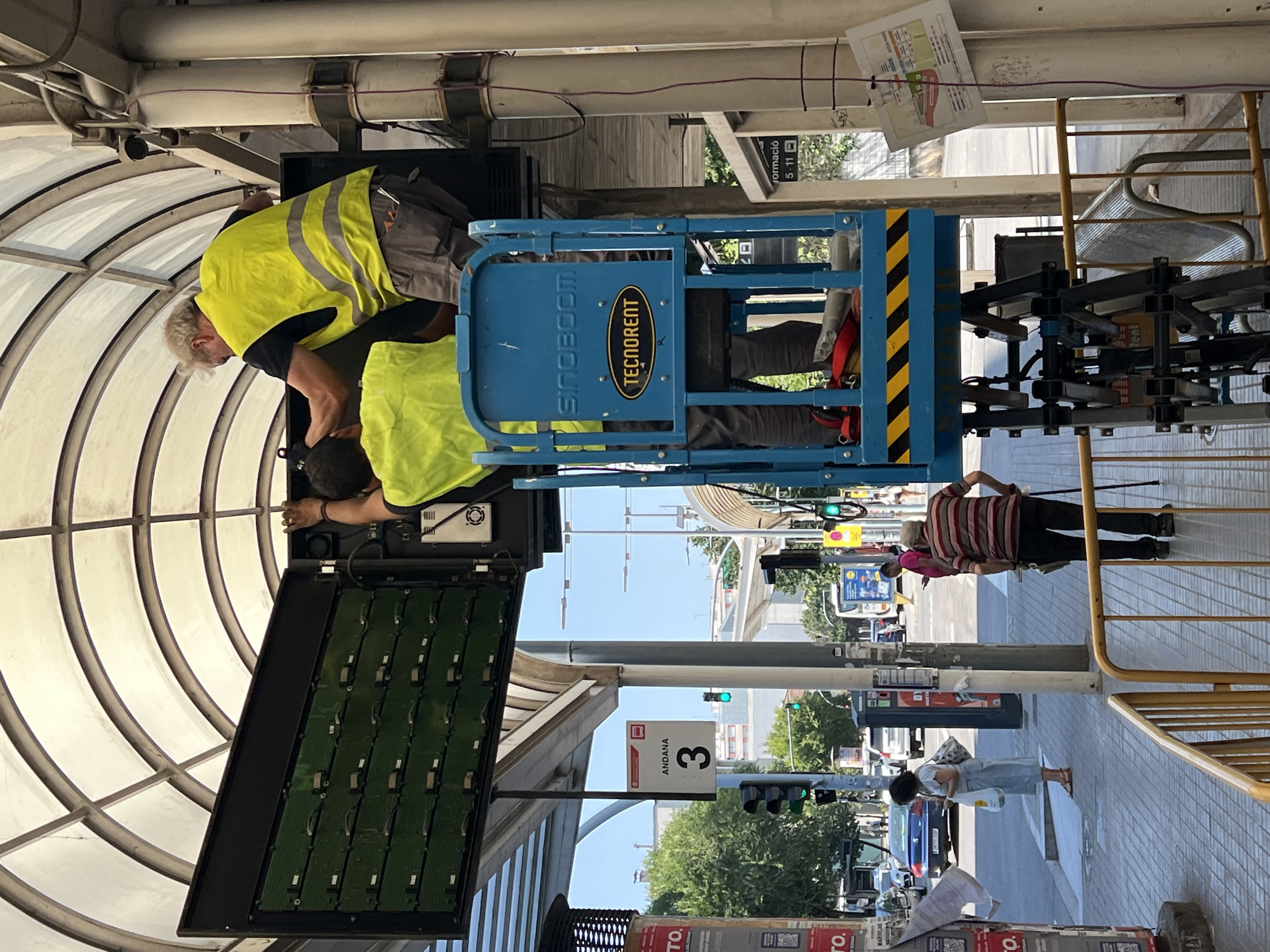 Two individuals, one wearing a high-visibility yellow vest, are working on an electronic display panel. The display panel is open, revealing rows of green circuit boards. They appear to be using a hydraulic lift, indicated by a blue platform labeled "TECNOCRENT" and "SINOBOOM". The scene takes place outdoors, under a bright, clear sky, suggesting daytime with good weather.
In the background, street elements are visible, including traffic lights, trees, and buildings. A sign with the number "3" and the word "ANDANA" is mounted on a pole. Other people are seen walking on the sidewalk, some carrying bags. A yellow metal fence is also present. The overall setting suggests an urban or semi-urban environment.
The activity appears to be maintenance or installation of the electronic display. The workers are focused on their task, and there are no immediate indicators of distress or urgency. The presence of the high-visibility vests and the lift suggests a professional work environment.
Visible text includes "TECNOCRENT", "SINOBOOM", "ANDANA", "3", and some unreadable text on posters in the background and on the electronic panel itself. The location context identifies the scene as Granollers, Spain.