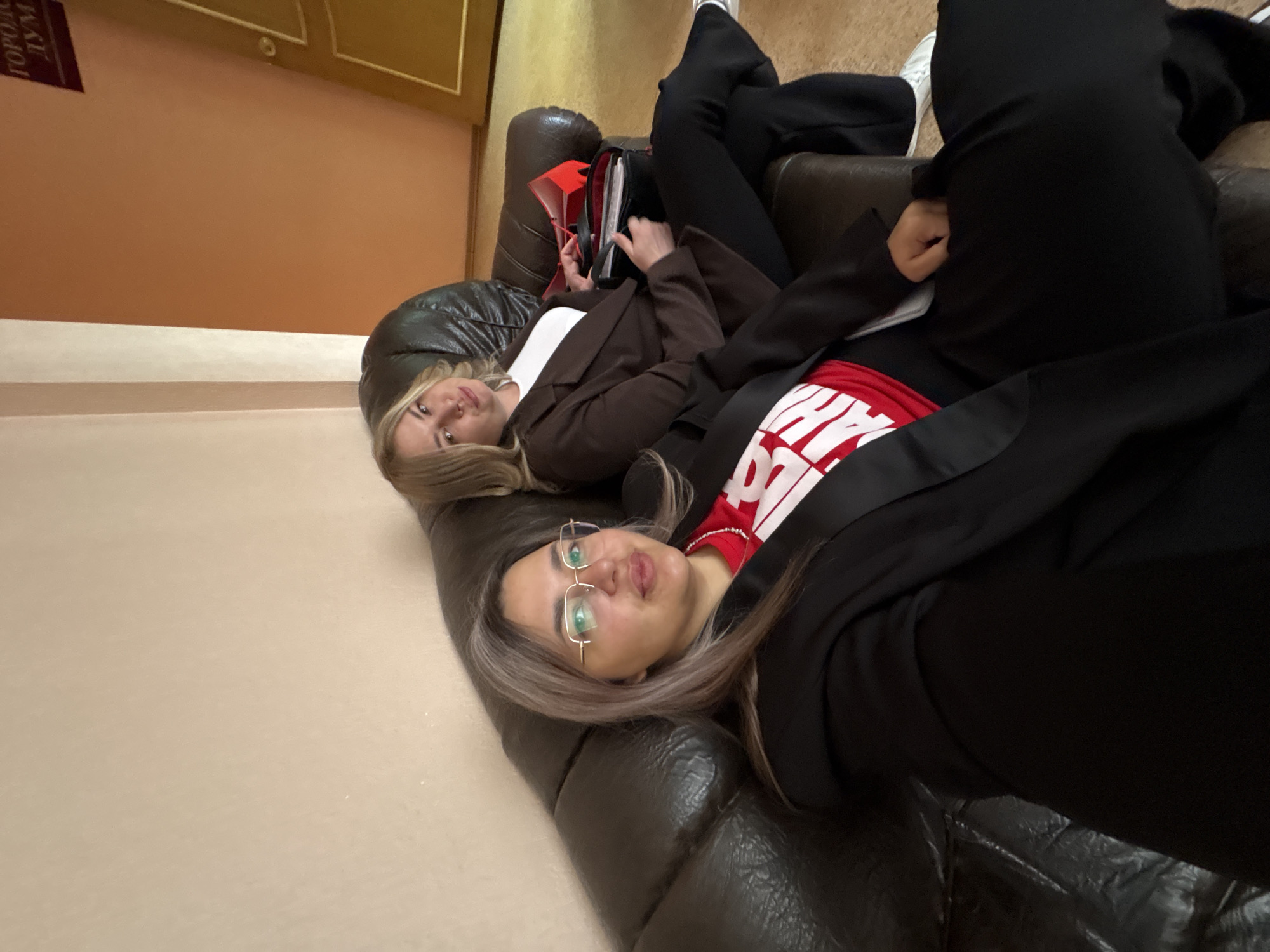 Two women are depicted resting on a dark leather sofa in what appears to be an office or waiting area in Birobidzhan, Russia. The image is taken from a low angle, with the subjects appearing to be lying down from the viewer's perspective. The woman in the foreground has light brown hair, is wearing glasses, and looks directly at the camera with a neutral expression. She is dressed in a black blazer over a red t-shirt with partially visible white text. Behind her, another woman with lighter hair is partially visible, wearing a brown blazer over a white t-shirt and dark pants. Her feet, clad in white sneakers, are visible raised on the armrest of the sofa. She is looking away from the camera, seemingly occupied with a red folder and a device in her hands.
The setting includes plain light brown walls and a door or panel with gold trim in the background. A sign is partially visible on the wall in the upper left, displaying Cyrillic text that reads "ГОРОДСК..." (Gorodsk...) and "ДУМА" (Duma), indicating a "City Council" or "City Duma." This textual cue, combined with the provided location context, suggests the scene is within a municipal government building in Birobidzhan. The overall atmosphere appears casual despite the formal attire, suggesting a moment of rest or waiting.