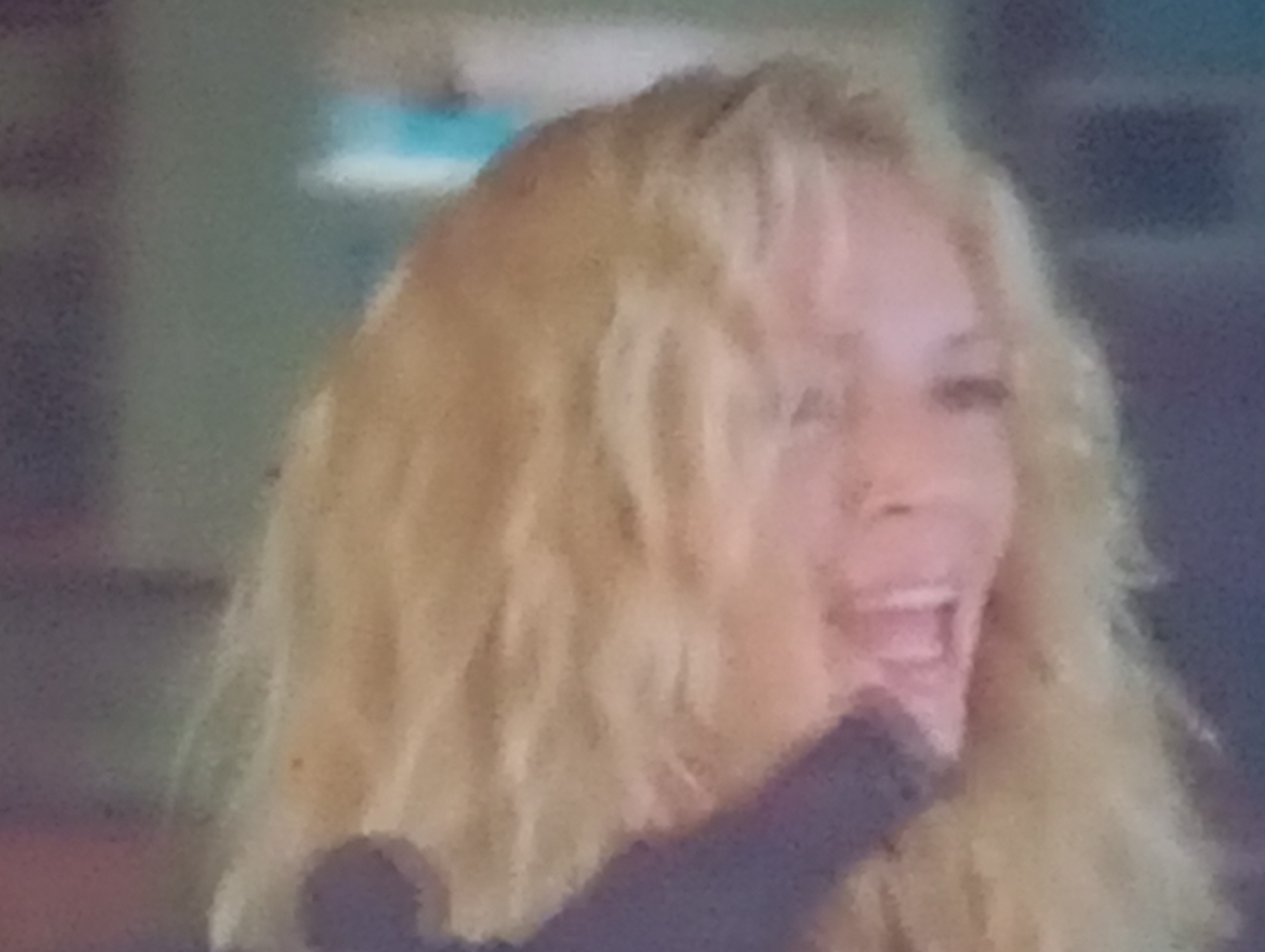 This is a close-up, somewhat blurry, and pixelated image of a woman with blonde, wavy hair. Her mouth is open as if she is laughing or speaking animatedly, and her eyes are not clearly visible. She appears to be wearing a dark-colored garment. The background is out of focus, showing a mix of muted colors, with hints of light blue and darker shades, suggesting an indoor setting. The lighting is somewhat dim. There are no visible text elements in the image. The overall impression is that of a candid moment, capturing an expression of joy or strong emotion.