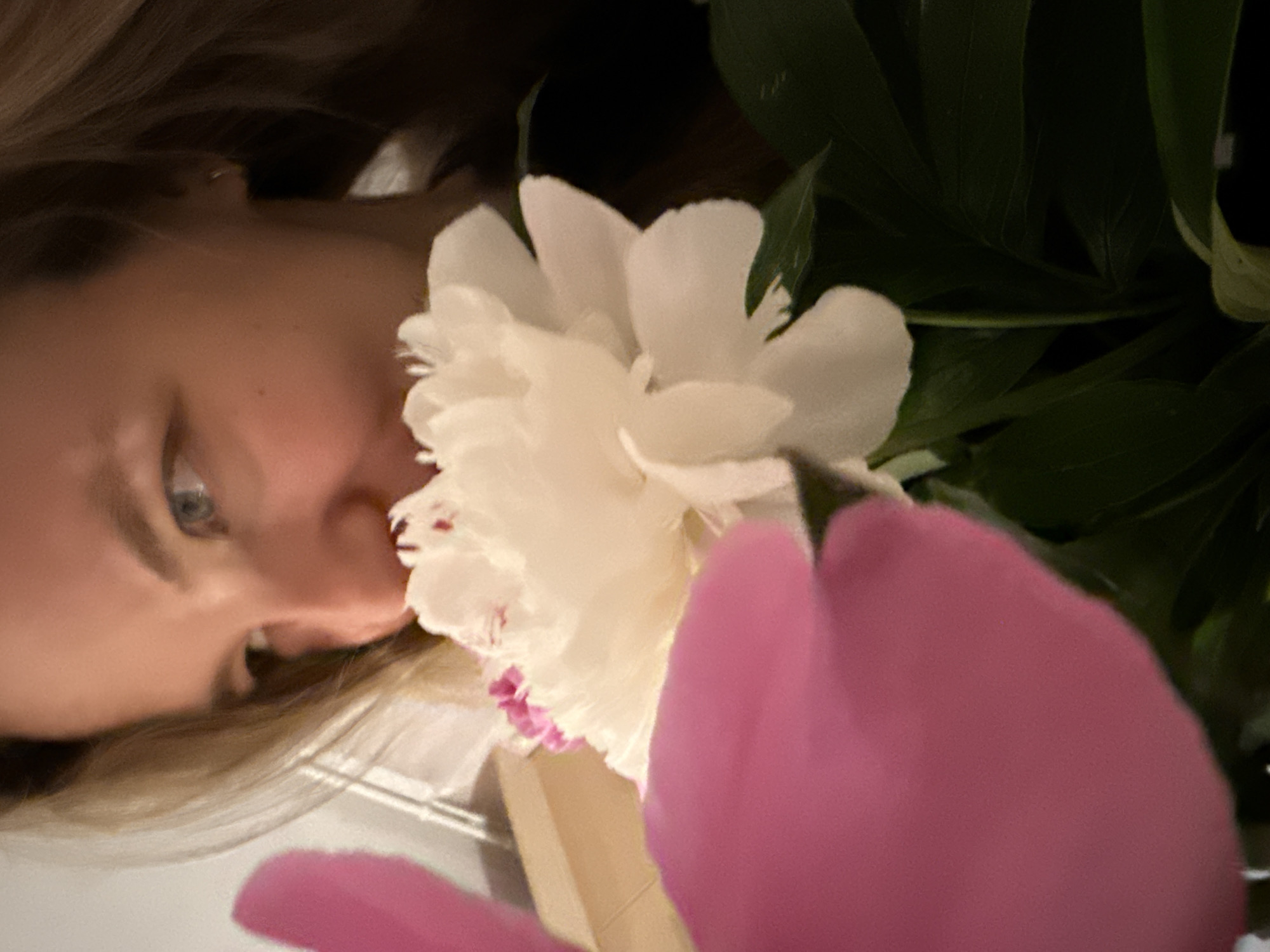 The image portrays an adult female engaged with a bouquet of flowers in an indoor setting. Her face is partially visible, featuring fair skin, light-colored eyes directed downwards and to the right, and long, light brown hair. A small, gold-colored hoop earring is discernible in her right earlobe. Her nose is deeply positioned among the petals of a prominent white peony, indicating the action of smelling the flower. This white peony exhibits delicate petals with subtle pink-red hues concentrated at its core. A large, out-of-focus bright pink peony occupies the immediate foreground on the left, partially obscuring the woman's left profile and other floral elements. Several dark green leaves are visible beneath the white peony, belonging to the bouquet. The background is blurred, depicting a light-toned interior wall with a faint vertical structural detail on the upper left. The visual information within the image does not provide any specific identifiers for an urban location such as Vladimir, Russia.