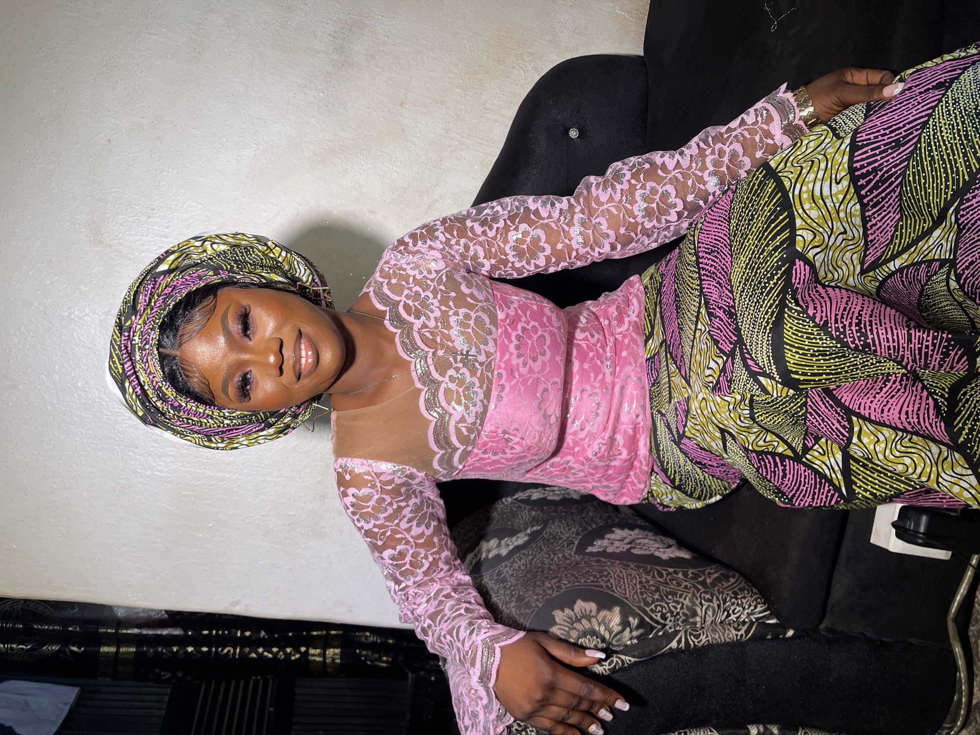 A young Black woman is depicted reclining on a dark sofa, looking towards the camera with a joyful smile. She is adorned in traditional Nigerian attire, featuring a vibrant headwrap (gele) and a matching wrapper or skirt, both patterned with striking yellow-green, pink, and black leaf-like designs. Her top is a bright pink, form-fitting garment with long, sheer lace sleeves detailed with silver embellishments and a nude-colored illusion neckline.
Her makeup is well-applied, highlighting her eyes with defined eyelashes and her lips with a glossy finish. She wears a thin silver chain necklace and a gold-toned bracelet on her right wrist, with her fingernails showcasing light-colored polish. The sofa is dark, likely black, and includes a patterned throw pillow. The background consists of a plain, light-colored wall that appears to have a subtle sparkle or texture. The setting is indoors, possibly a living room, suggesting a relaxed and personal atmosphere. The woman's confident and happy expression conveys a sense of celebration or contentment. The image captures cultural elegance and beauty, originating from Akwanga, Nigeria. There is no visible text in the image.