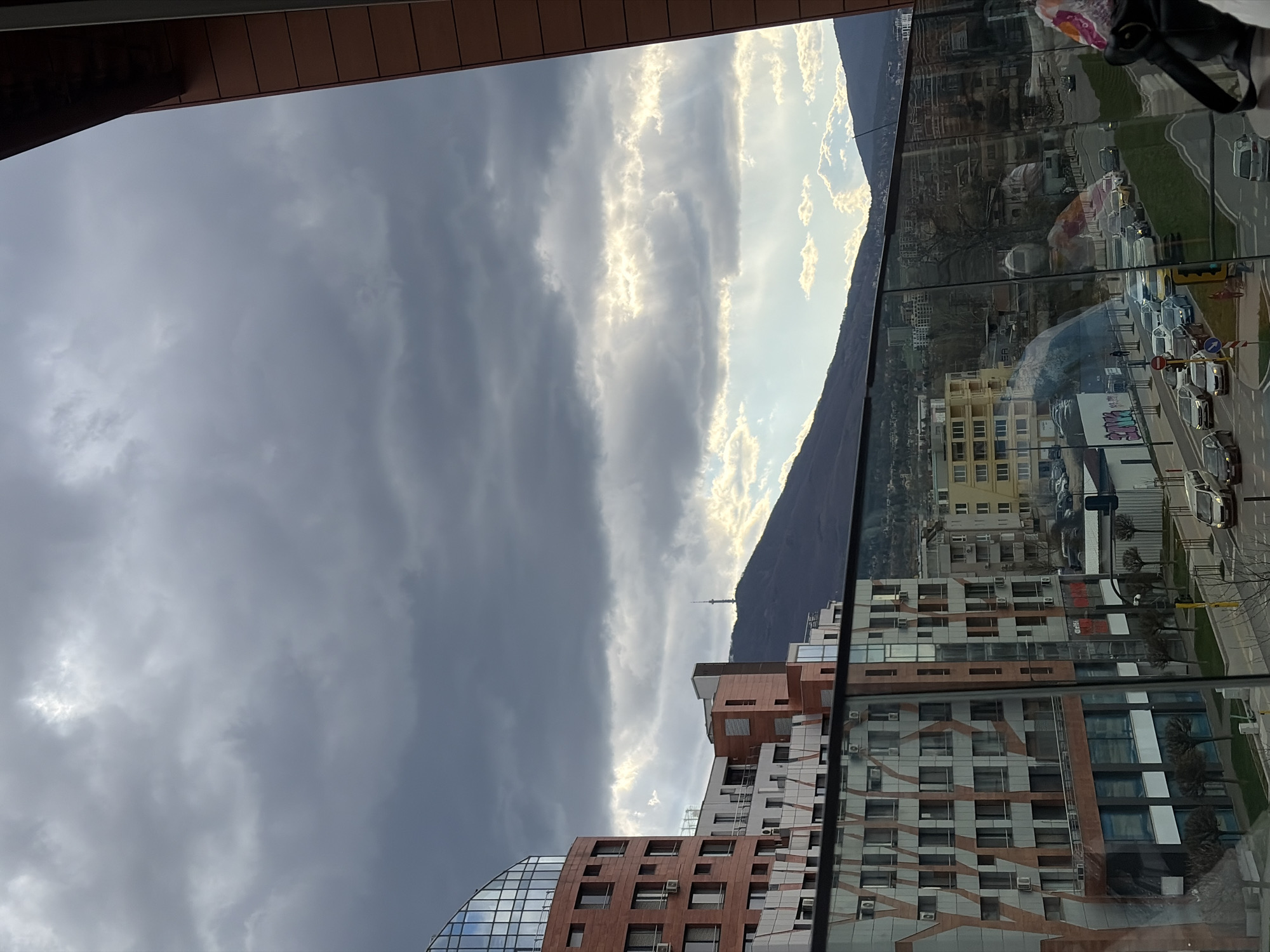 The image is taken from within a building in Sofia, Bulgaria, looking out through a window. The view includes the city's architecture, a mountain range, and the sky. The foreground features parts of the building from which the photo is taken, including what appears to be a window frame. The architecture in the city below consists of modern buildings, including high-rise structures with glass and brick facades. In the distance, a mountain range is visible, silhouetted against a cloudy sky. The clouds are varied, with some appearing dark and stormy, while others are illuminated by sunlight. The image also reflects the surrounding environment, creating a layered effect. The reflection on the glass window also shows the opposite buildings, vehicles, and streetlights.