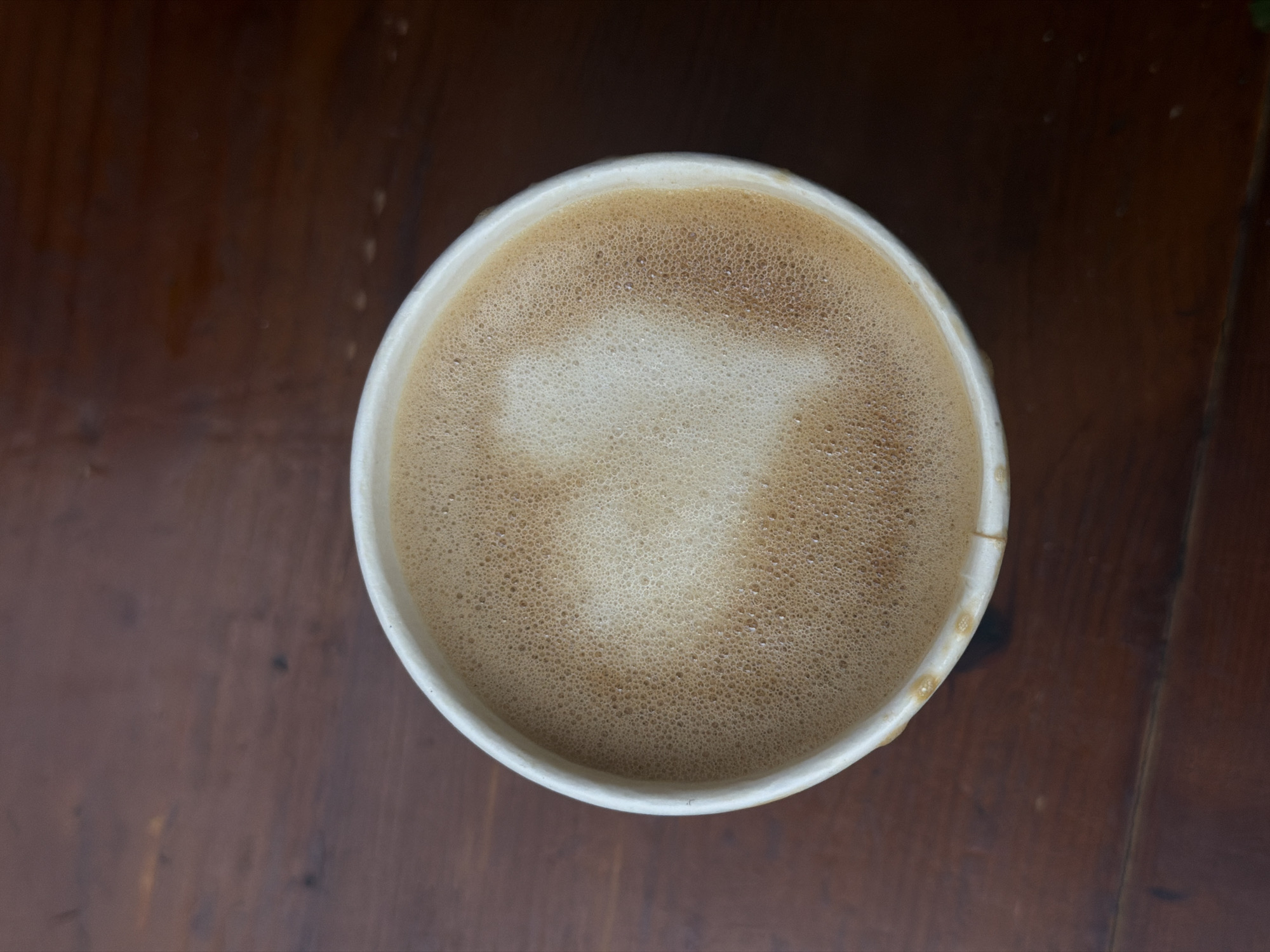 An overhead shot captures a single white paper cup filled with a frothy coffee beverage, likely a latte or cappuccino, resting on a dark brown wooden surface. The surface of the coffee is covered in a dense layer of foamed milk, with a lighter, indistinct, perhaps heart-shaped pattern at its center, surrounded by numerous small bubbles. The rim of the white cup is slightly visible, showing subtle marks of use or texture. The wooden table beneath has a visible grain and appears to have a few faint water marks or specks, suggesting a natural and perhaps well-used setting. No people are visible, and the focus is entirely on the coffee. The lighting appears soft and even, suggesting an indoor or shaded outdoor environment. No text is visible on the cup or the table. The image evokes a calm and simple moment of enjoying a beverage, potentially in a cafe or at home in Đà Nẵng, Vietnam.