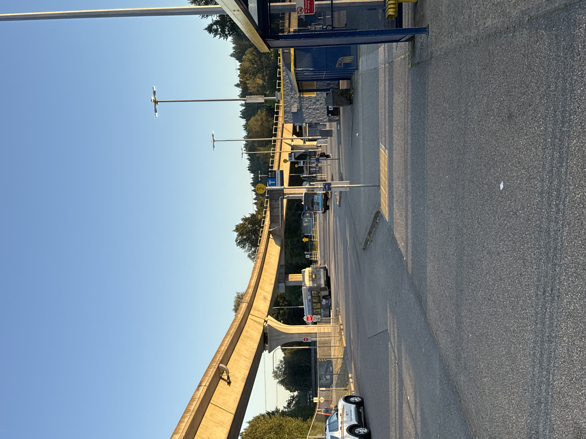 The image captures an outdoor scene at a bus loop or transit station, likely during the late afternoon or early evening due to the warm lighting and long shadows. The location is identified as Coquitlam, Canada, suggested by the presence of TransLink infrastructure.
Dominating the upper portion of the image is an elevated concrete guideway, characteristic of SkyTrain tracks in the Vancouver area, specifically the Expo Line that serves Coquitlam. The guideway is supported by large concrete pillars.
In the foreground, a paved road surface shows distinct lane markings and a pedestrian crossing with tactile paving. To the right, a bus shelter with a yellow roof and blue supporting posts is visible. A "No Smoking or Vaping" sign is affixed to the shelter. In front of the shelter are yellow benches.
Several buses are present. One bus, a standard transit bus with a route display, is parked at what appears to be a bus bay labeled "Bay 2." Another, older-looking bus with a grey exterior, is also visible. A white security or transit patrol vehicle with text indicating "Transit Supervision" is parked on the left side of the road.
The environment includes mature trees lining the background, suggesting a suburban or park-like setting. Streetlights on tall poles are also prominent, equipped with multiple lamps. The sky is clear and blue. Scattered trash receptacles are also visible near the bus shelter. One person, dressed in dark clothing, is walking in the distance near the bus bays. A stop sign is visible on a pole near the elevated guideway.