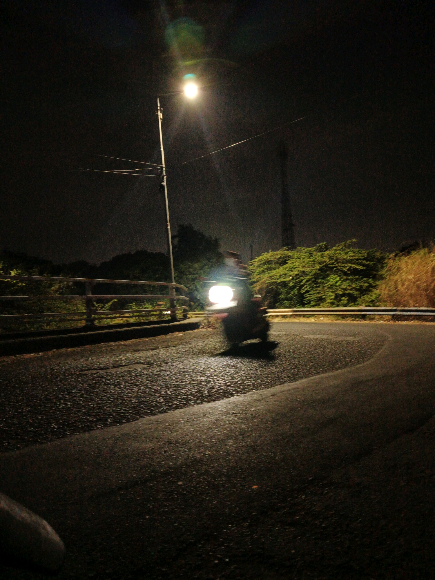 The image depicts a night scene on a wet, winding road in Kludan, Indonesia. A person is riding a scooter, illuminated by the scooter's headlight and a nearby streetlamp. The road surface is asphalt, appearing dark and reflective due to the moisture. On the left side of the road, there is a metal guardrail bordering the street. Beyond the guardrail, lush green vegetation and trees are visible. On the right side of the road, dry, tall grass lines the embankment. The sky is dark, with a bright light source, likely a streetlamp, creating a halo effect. In the background, a tall, slender structure, possibly a utility pole or tower, is faintly visible against the dark sky. The overall impression is a dimly lit, atmospheric night drive.