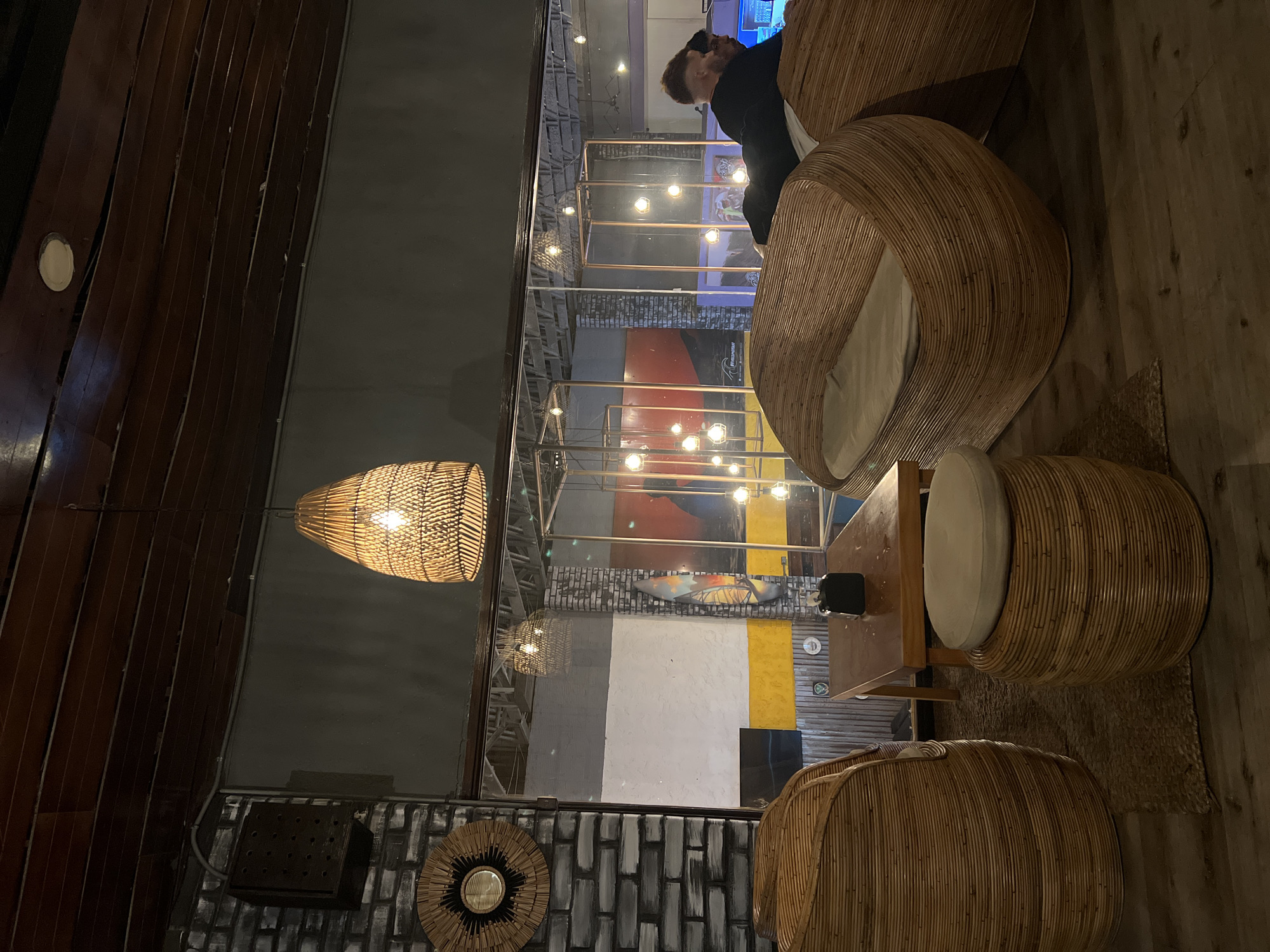 The image depicts an indoor scene within a commercial establishment in Arica, Chile, characterized by a modern, natural aesthetic and dim, warm lighting.
In the immediate foreground, dark wood plank flooring is visible, partially covered by a woven natural fiber rug. A cylindrical rattan stool with a cream cushion rests on the rug. To the left, a large, curved, open-weave rattan armchair with a cream cushion is positioned. A dark-stained rectangular wooden table, bearing a black square-shaped object, stands between this armchair and the stool. To the right, a second identical rattan armchair/daybed is present, with a cream cushion.
Two adult males are occupying the rattan armchair on the right. The prominent male, positioned centrally, has fair skin, short light-colored hair, and a beard. He wears a dark long-sleeved shirt and is leaning forward. A second male, with darker hair and skin, is partially obscured behind and to the right of the first, also wearing a dark shirt. Both individuals are looking downwards, suggesting engagement with an object or activity below their field of view.
The ceiling consists of dark wooden slats, featuring recessed circular lights and a central hanging rattan pendant lamp with an exposed incandescent bulb. To the left, a grey-painted brick wall displays a circular rattan wall hanging with a central mirror. A large, framed glass partition separates the foreground seating area from a more expansive background space.
Beyond the glass partition, the background reveals additional sections of brick wall, some painted with horizontal grey, white, and yellow stripes, and others featuring a large mural with dominant red, orange, and dark tones, including what appears to be an abstract surfboard graphic. Multiple geometric copper-toned metal frames are visible, interspersed with numerous suspended spherical light fixtures. Reflections of the interior lighting are present on the glass partition.