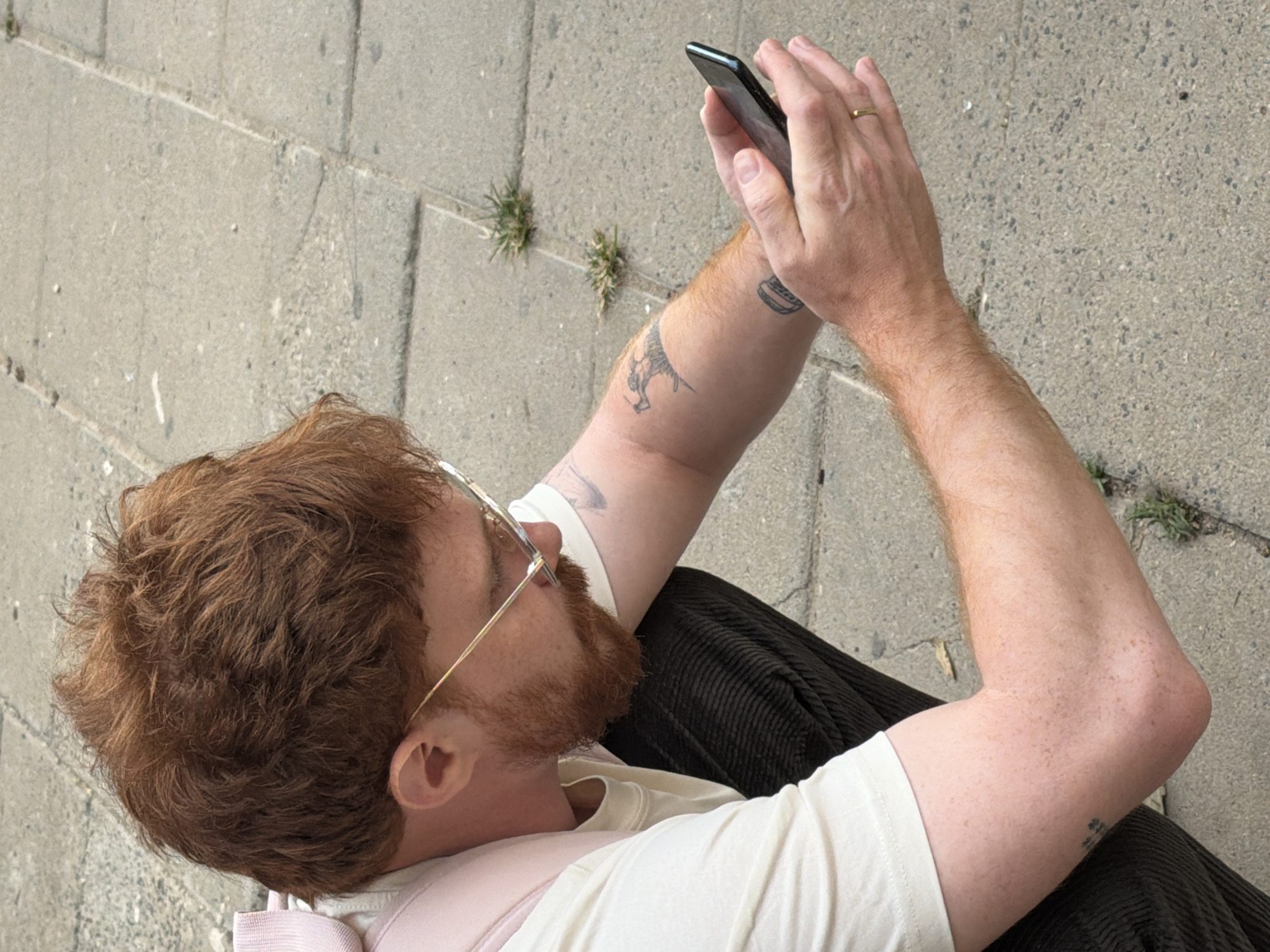 From an elevated perspective, a person is seen using a mobile phone while sitting on a concrete surface. The person has red hair and a beard, is wearing glasses, a white t-shirt with a pink under layer, and dark-colored corduroy pants. The person's left arm is raised, holding the phone, with visible tattoos on the left arm. A gold ring is visible on the ring finger. The setting appears to be an outdoor urban environment, evidenced by the concrete ground with small patches of grass. The location in the city of Tel-Aviv, Israel, cannot be confirmed from the image alone.